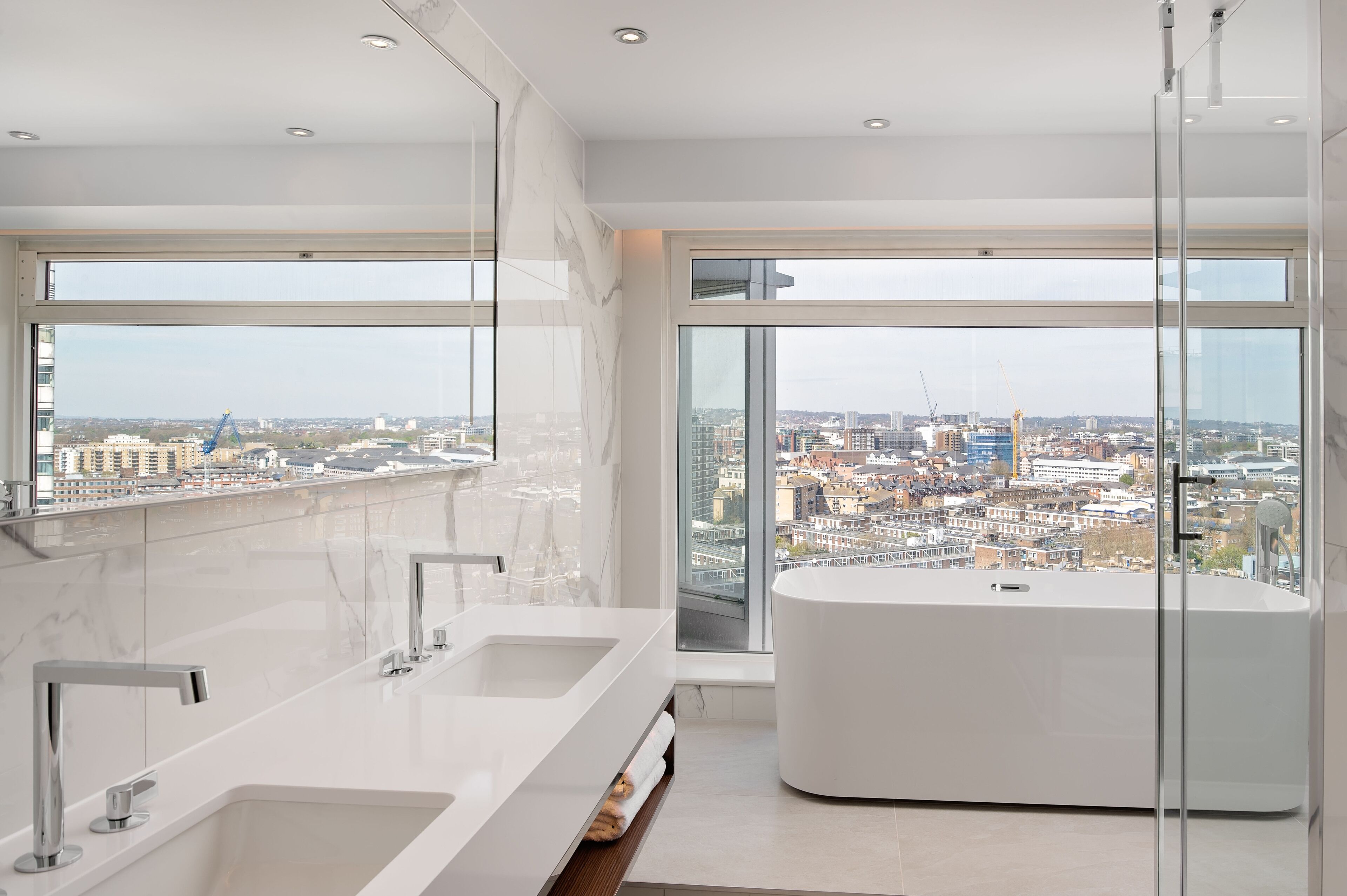 A modern bathroom with dual sinks, a large bathtub, and floor-to-ceiling windows offering a city view.