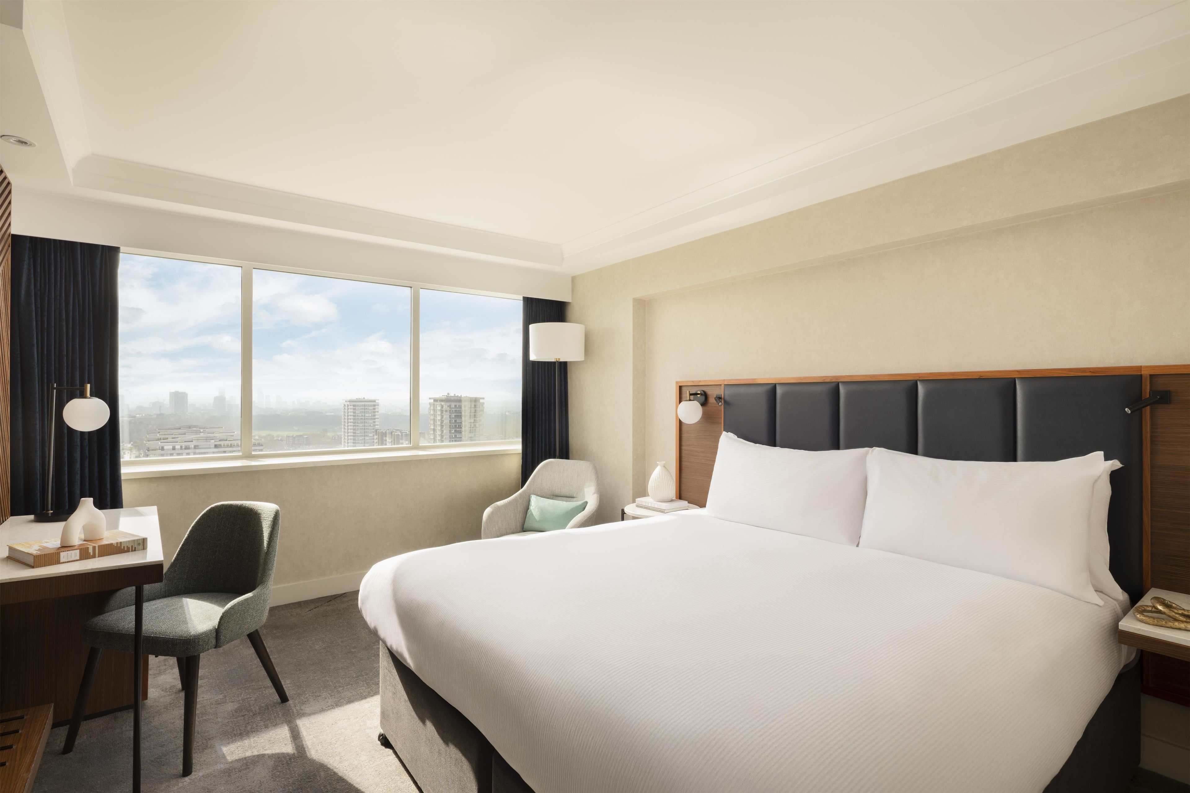 Ein Hotelzimmer mit einem großen Bett, einem Schreibtisch mit Stuhl, einem Fenster mit Stadtblick und einem Nachttisch mit Lampe.