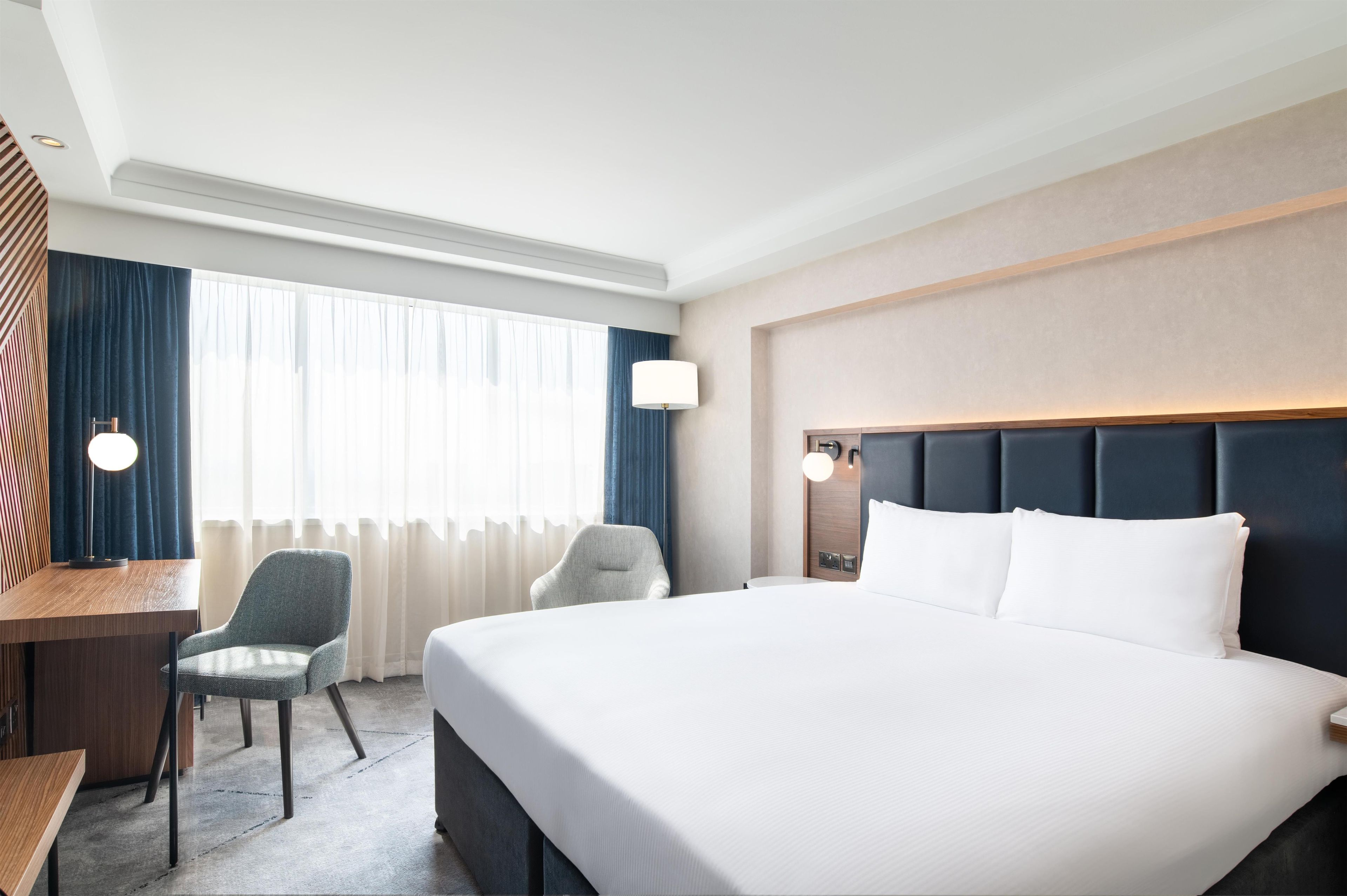 A hotel room with a large bed, a desk, two chairs, a lamp, and a view of the outdoors.
