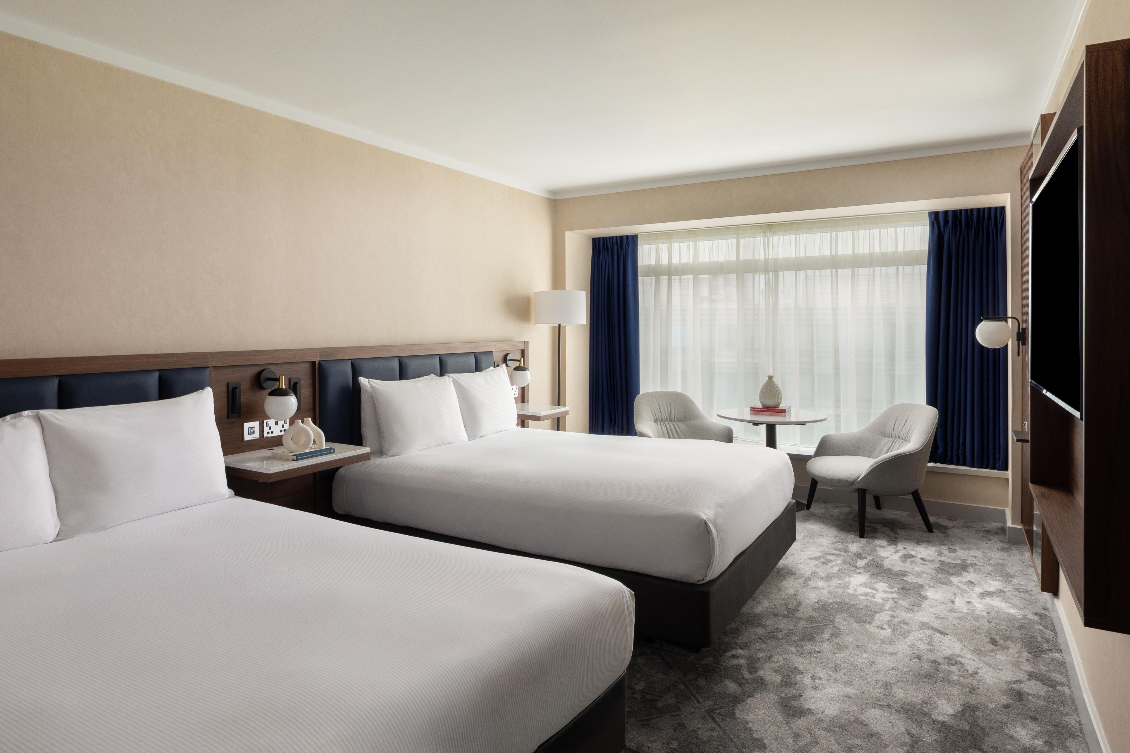 A hotel room with two beds, a desk, a chair, a lamp, and a view of the outdoors.