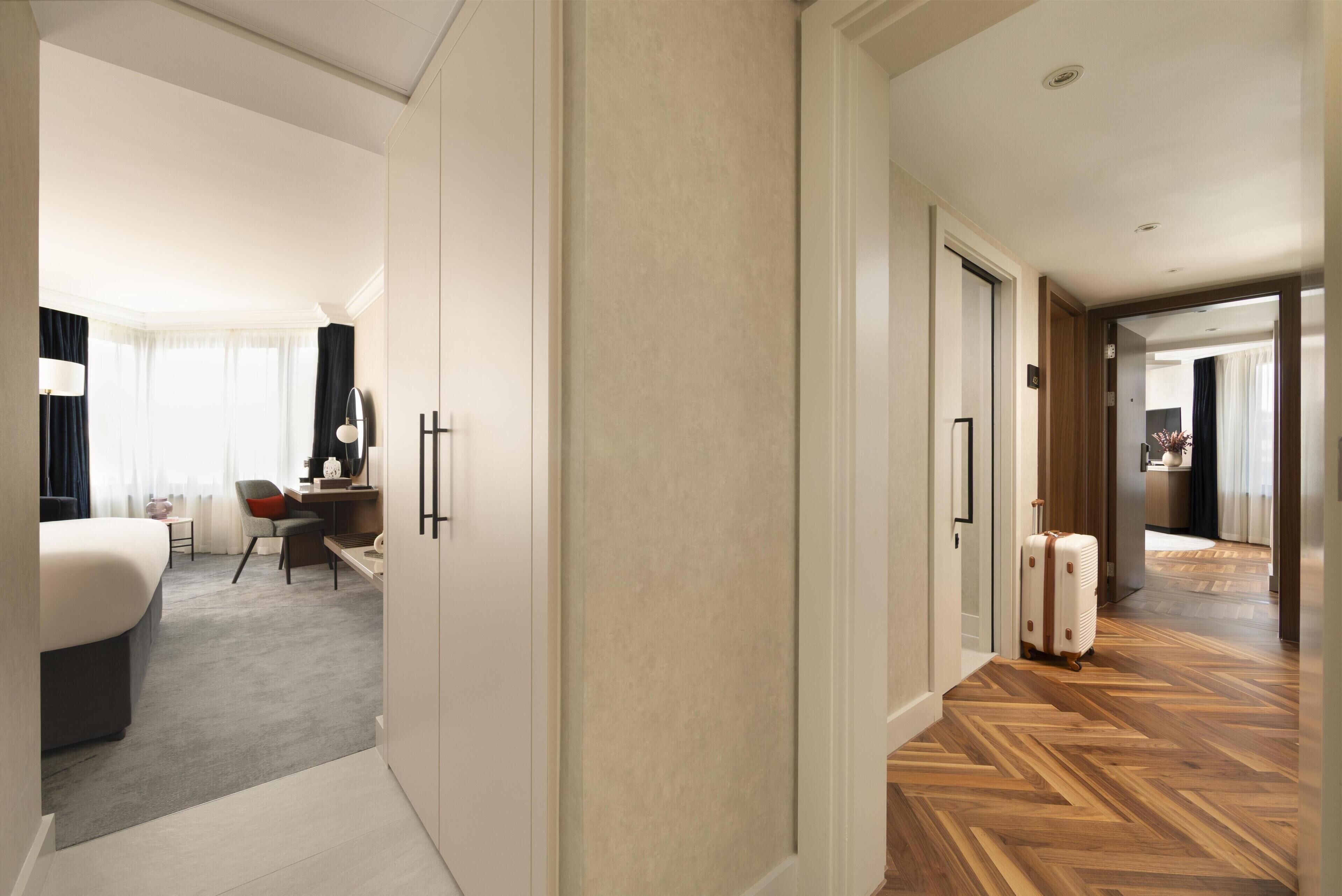 A hotel room with a bed, desk, chair, and a suitcase.