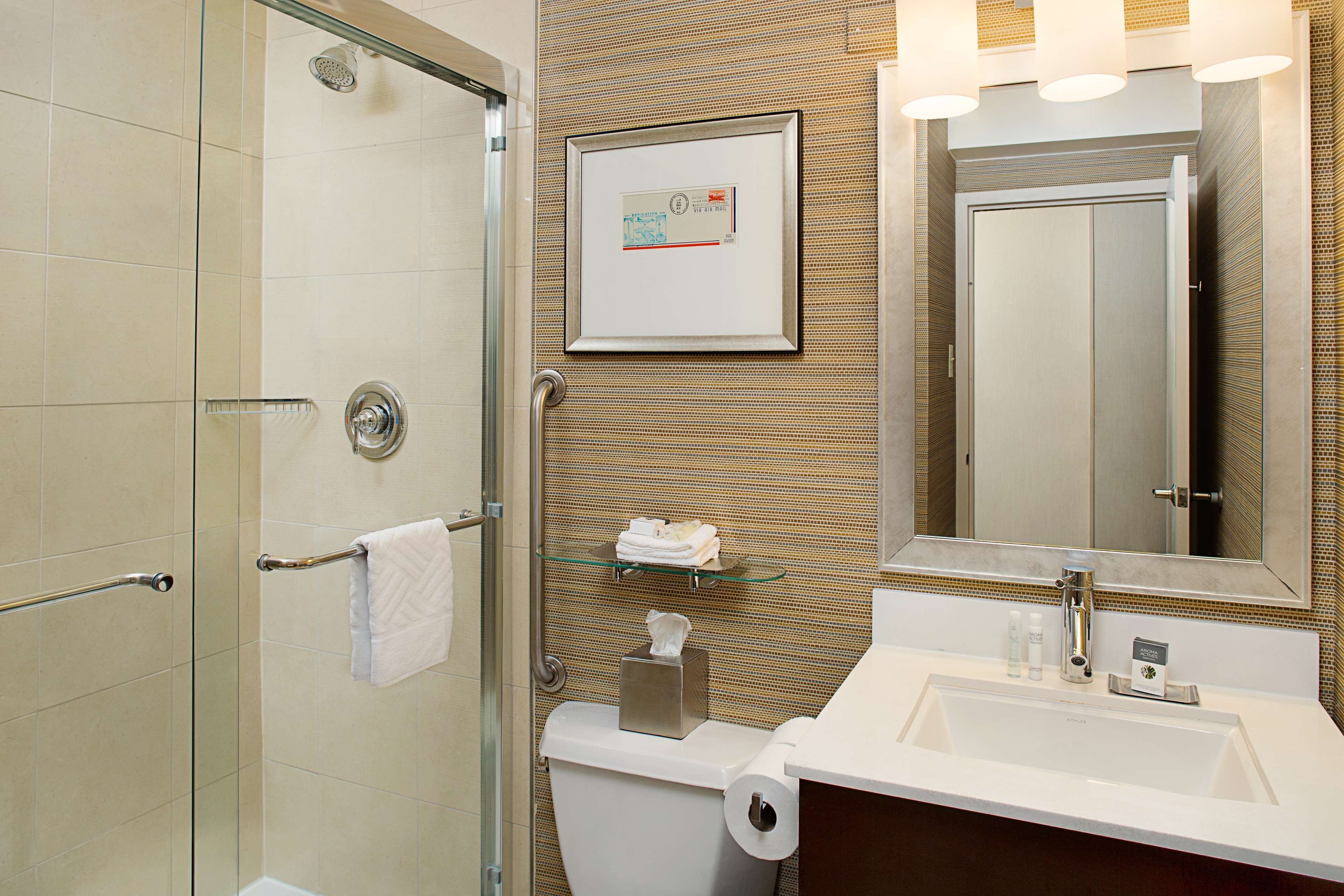 Un baño moderno con cabina de ducha de vidrio, un inodoro con soporte para toallas de papel, un lavabo con espejo arriba y un certificado enmarcado en la pared.