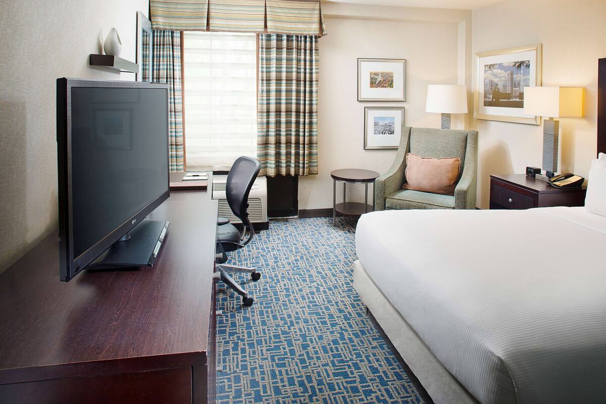 A hotel room with a large bed, a desk with a TV, a chair, a small table, and a lamp.