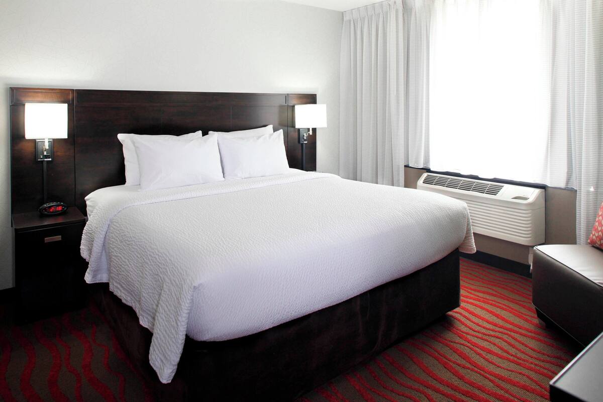 A hotel room with a large bed, two bedside lamps, a nightstand, a red patterned carpet, and a window with curtains.