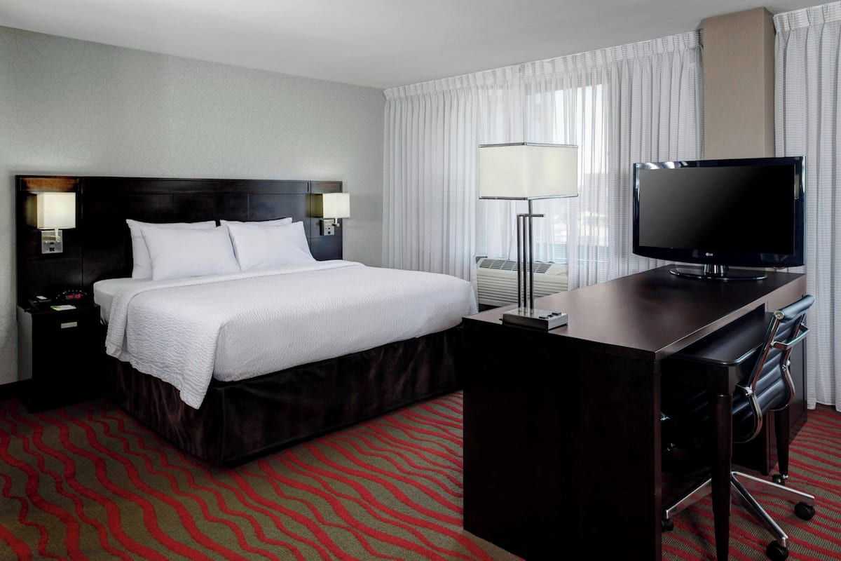 A hotel room with a large bed, a desk with a flat-screen TV, a lamp, and a red carpet.