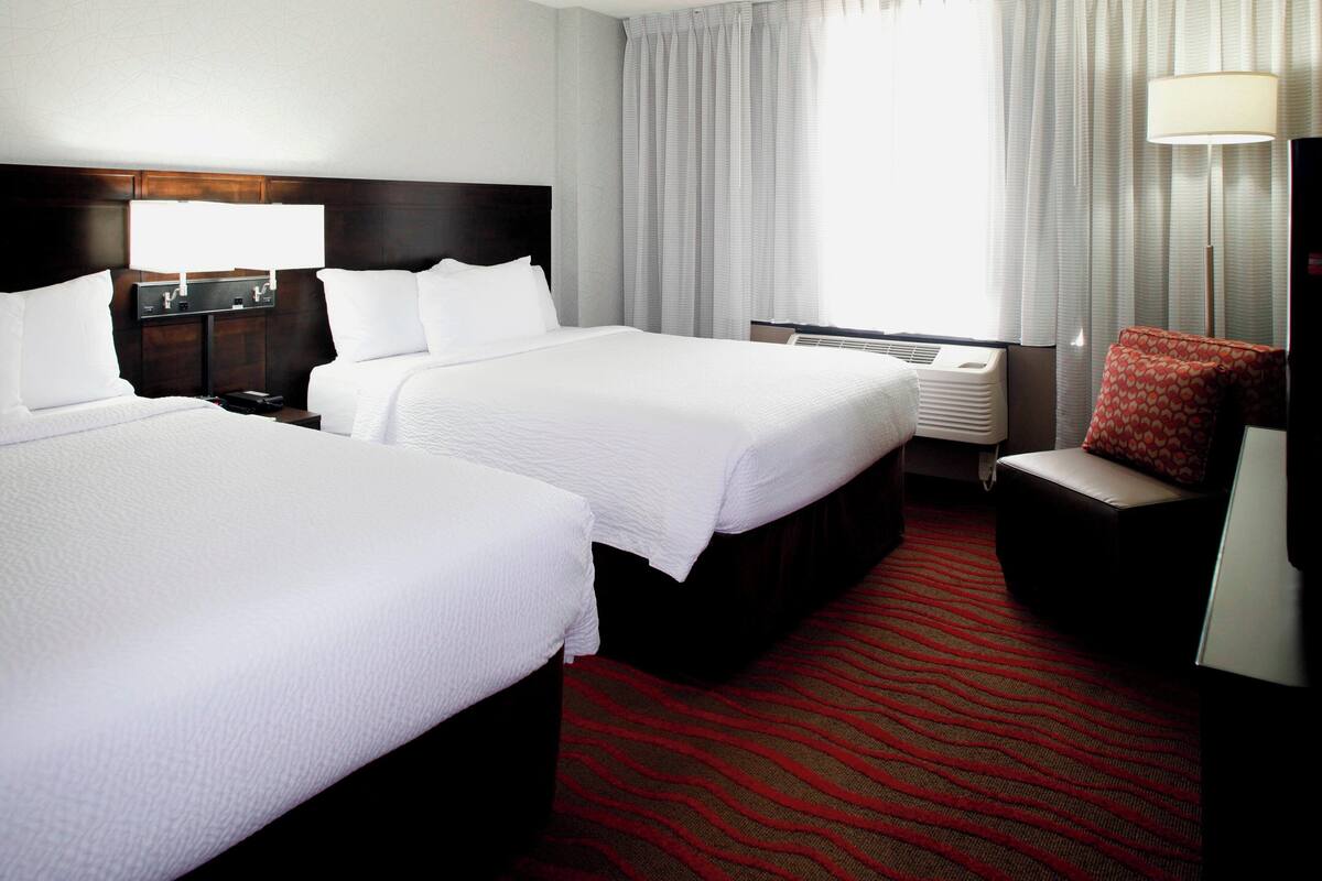 A hotel room with two beds, a red carpet, a nightstand with a lamp, and a red chair.