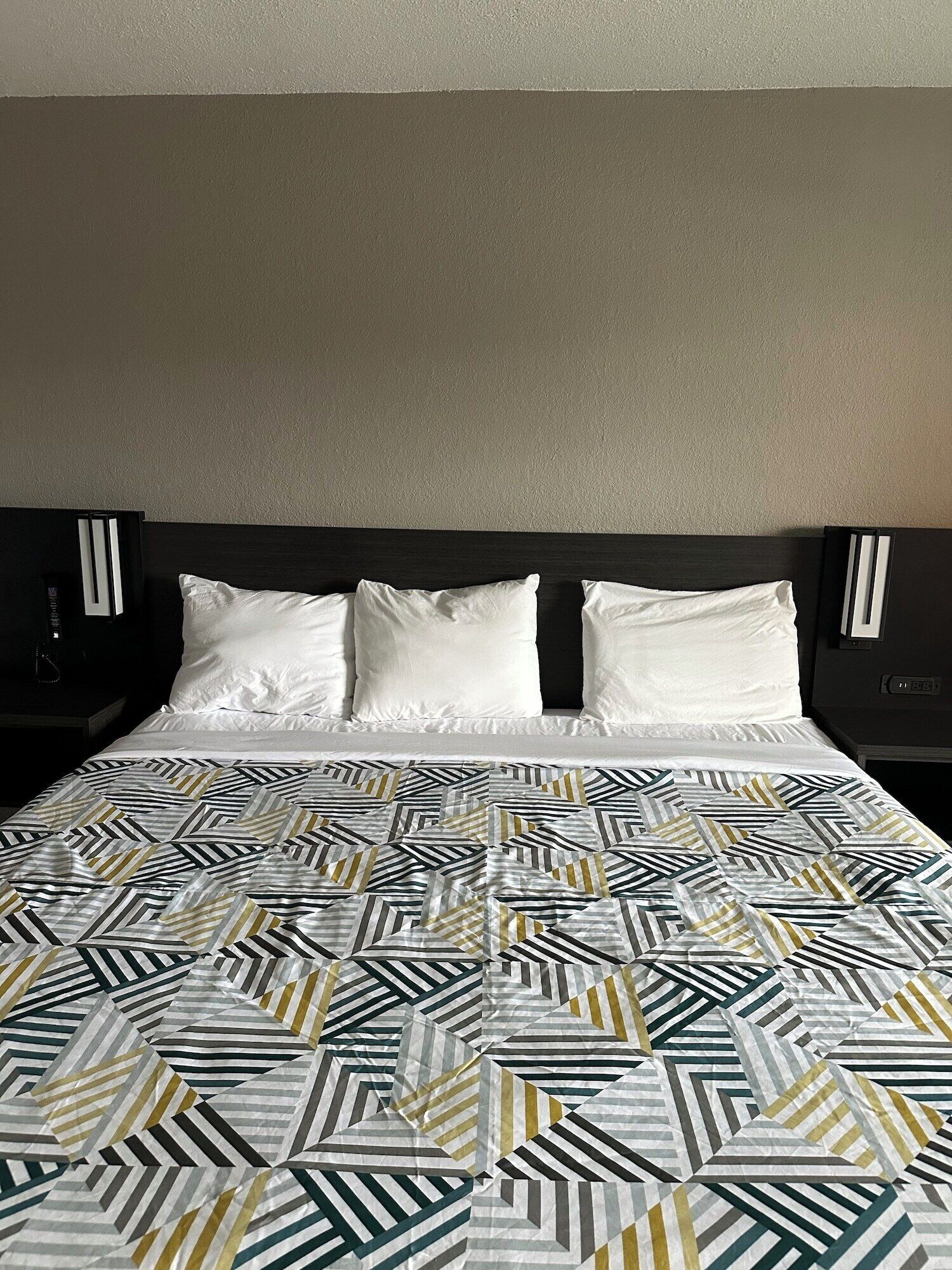 A hotel room with a bed, white pillows, and a geometric patterned bedspread.