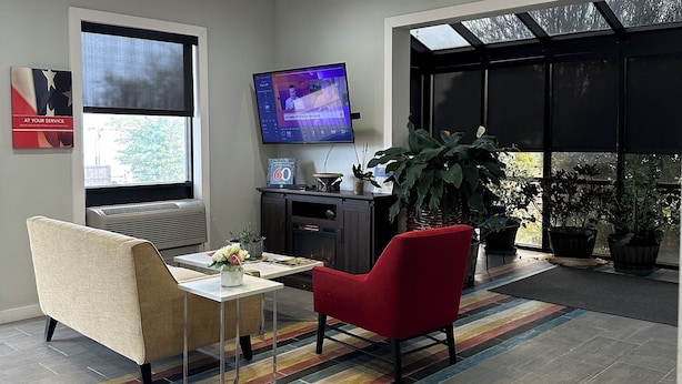 A modern living room with a flat-screen TV, a sofa, a red chair, a coffee table, and a large window with a view. - Motel 6 Brookhaven, MS (Brookhaven)