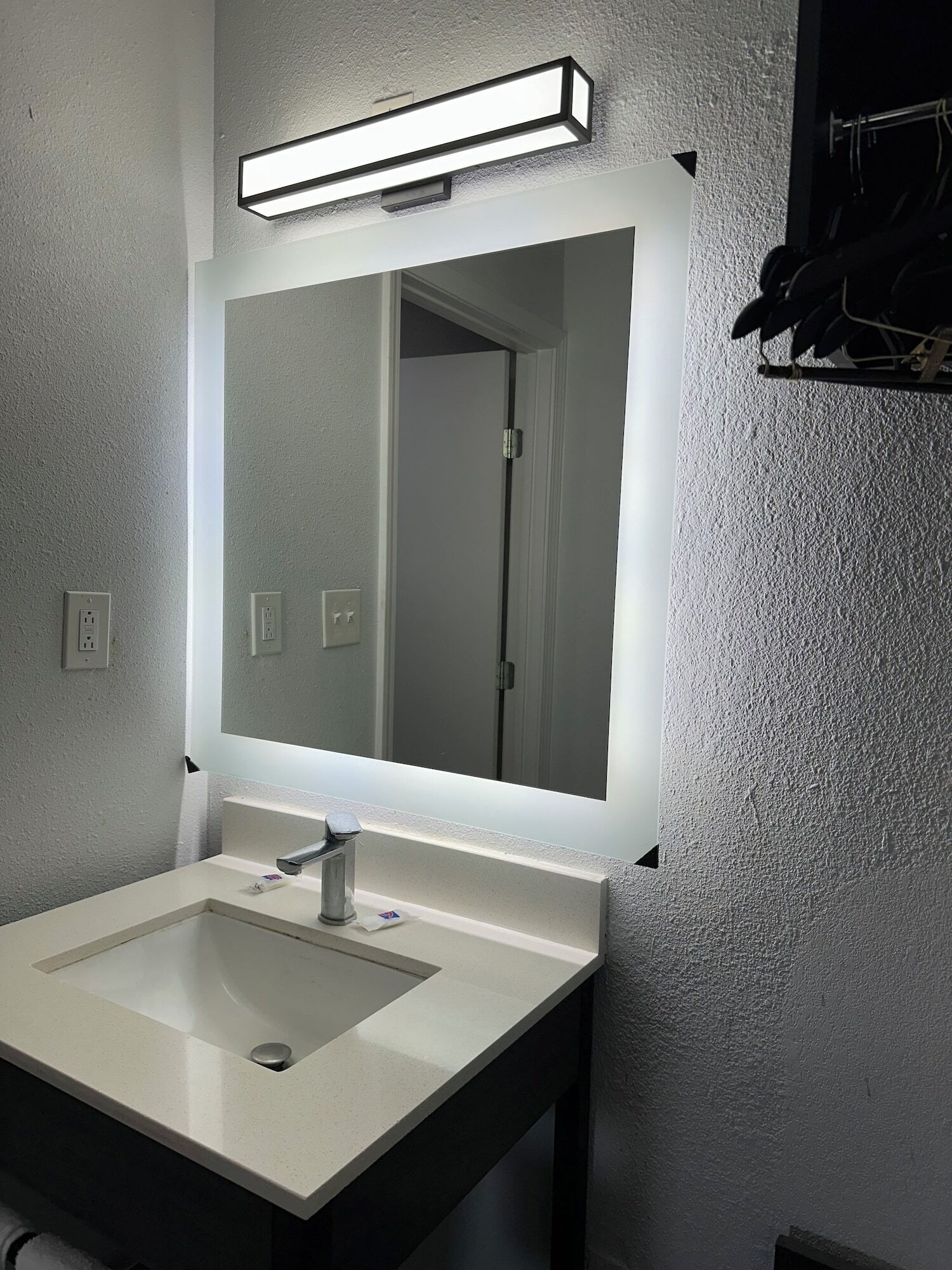 A bathroom with a rectangular sink, a mirror with a light above it, and a towel rack on the wall.