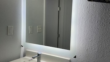 A bathroom with a rectangular sink, a mirror with a light above it, and a towel rack on the wall.