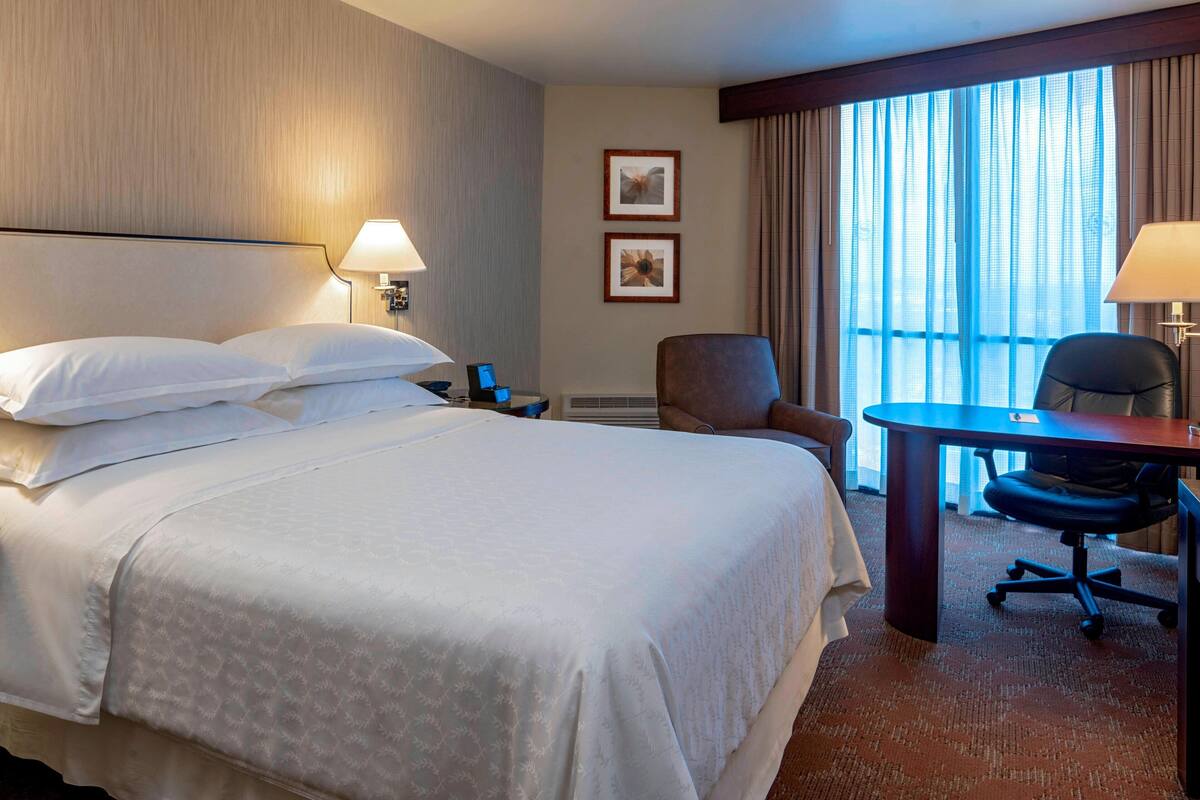 A hotel room with a large bed, two bedside lamps, a desk with a chair, and a window with curtains.