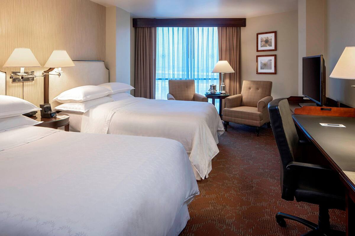 A hotel room with two beds, a desk, a chair, a TV, and a window with curtains.