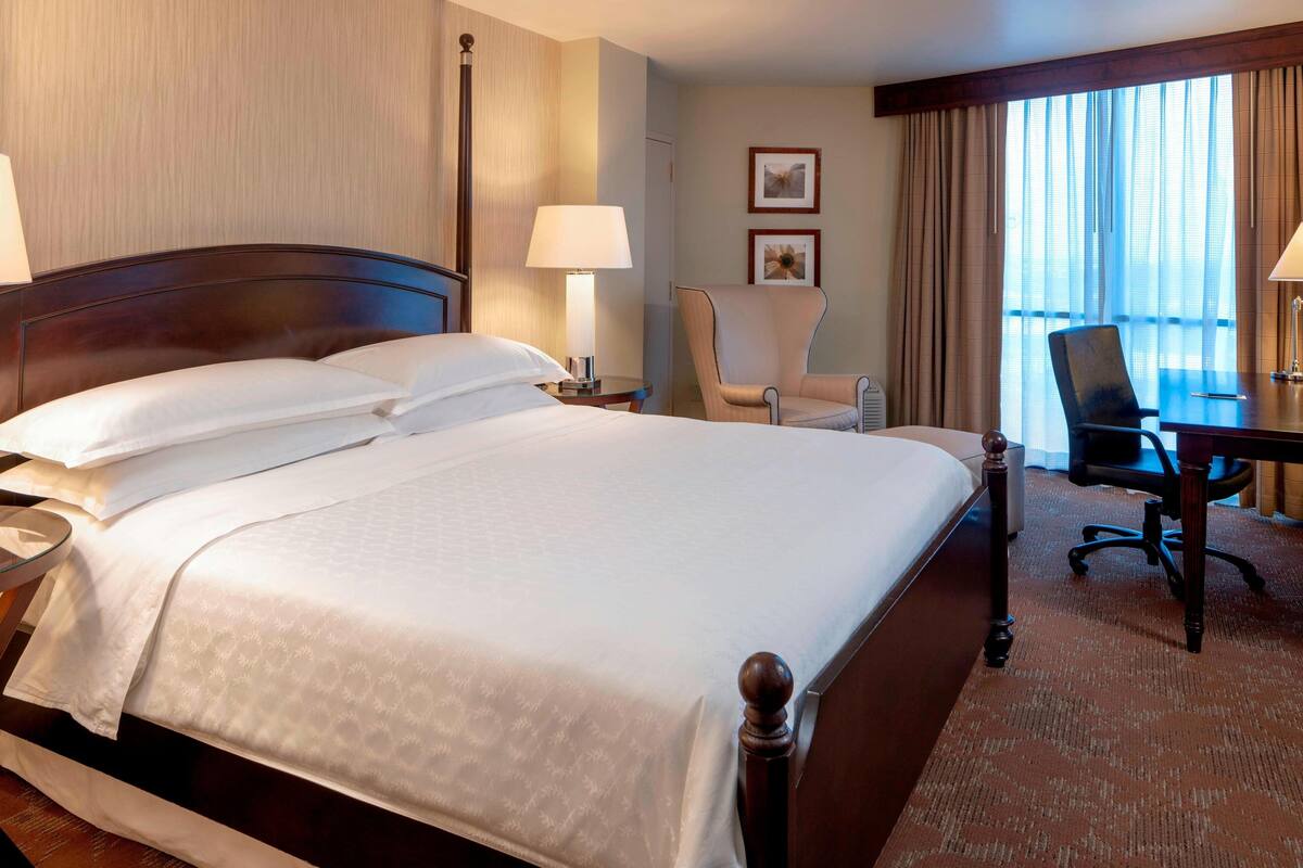 A hotel room with a large bed, a desk, a chair, and a window with curtains.