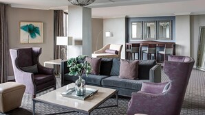 A modern hotel room with a living area featuring a sofa, armchairs, a coffee table, and a dining table.
