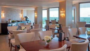 A dining area with a wooden table and white chairs, a kitchen in the background, and large windows offering a view of the outdoors.