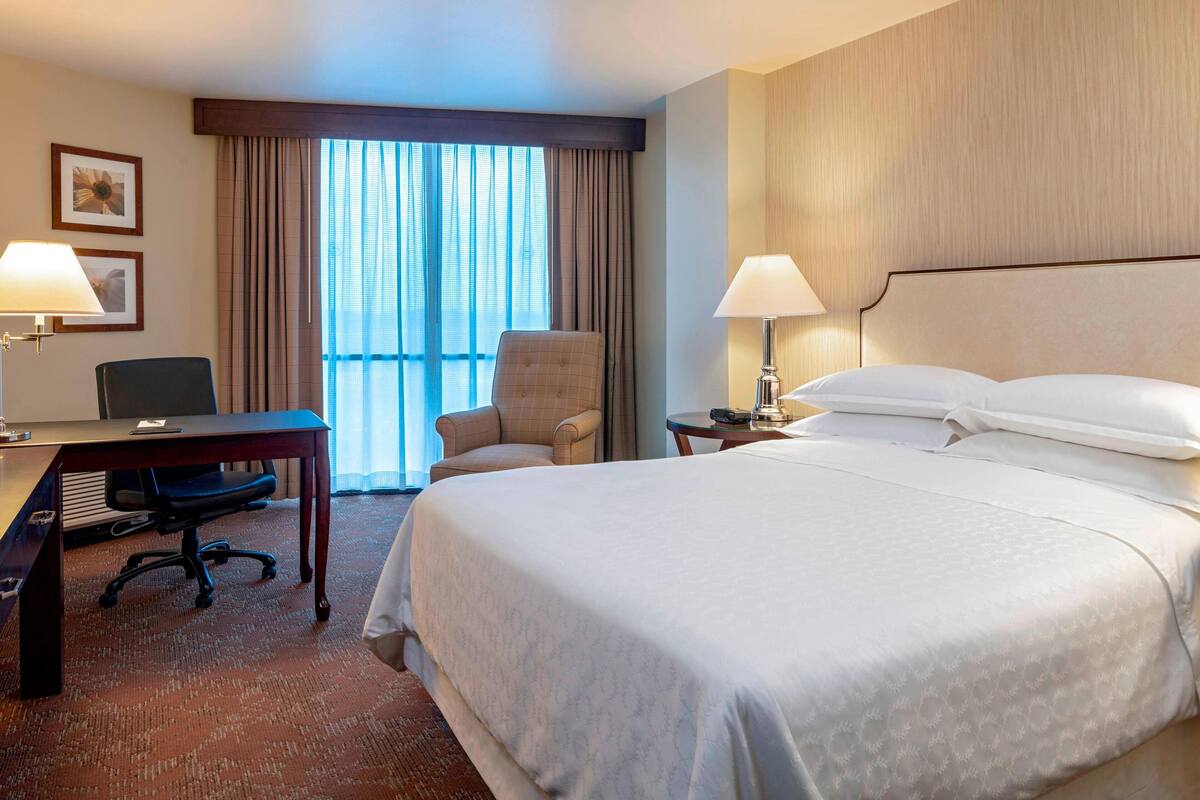 A hotel room with a large bed, a desk with a chair, a lamp, and a window with curtains.