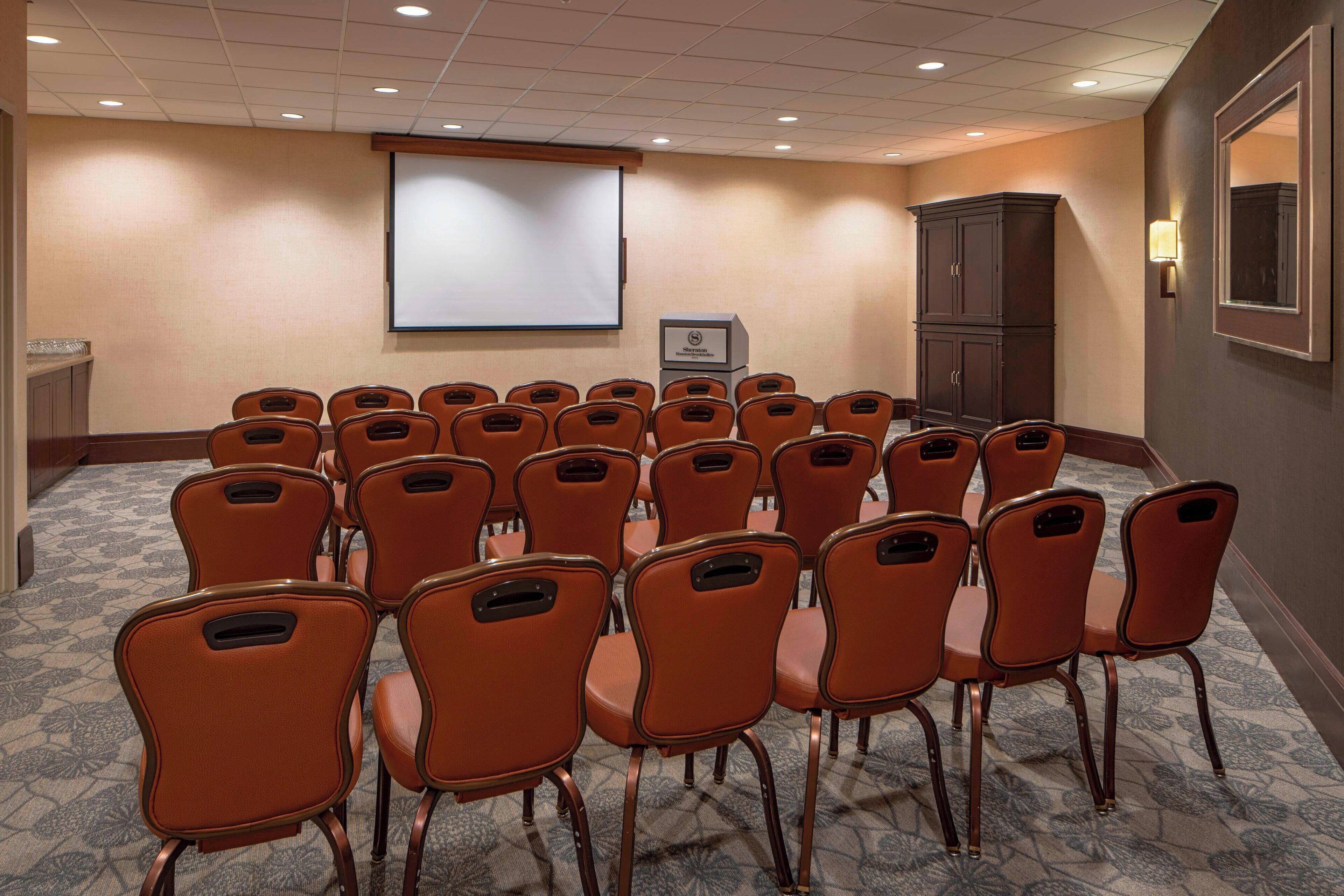 A conference room with rows of orange chairs, a projector screen, and a cabinet.