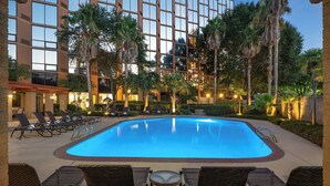 A swimming pool with lounge chairs and a table, surrounded by palm trees and a building with large windows.