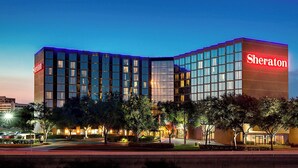 A Sheraton hotel with a modern glass facade, multiple floors, and a well-lit entrance area with trees and landscaping.