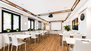 Une salle de conférence moderne, dotée de tables et de chaises blanches, d’un projecteur, d’un tableau blanc et d’une grande fenêtre donnant sur des plantes.