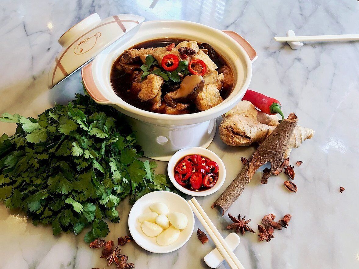 A bowl of soup with various ingredients and garnishes, accompanied by fresh herbs, chili peppers, and spices on a marble surface.