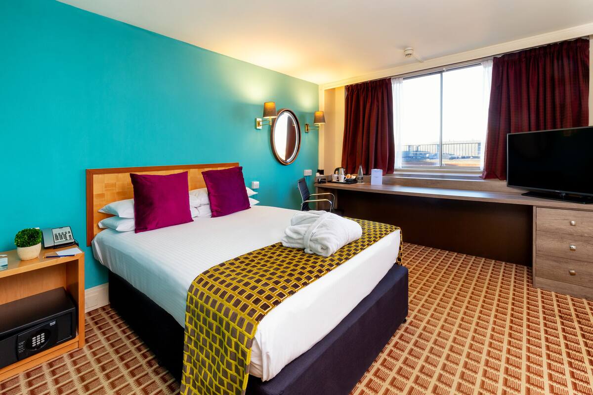 Ein Hotelzimmer mit Bett, Schreibtisch, Fernseher, einem Fenster mit Vorhängen und Blick ins Freie.