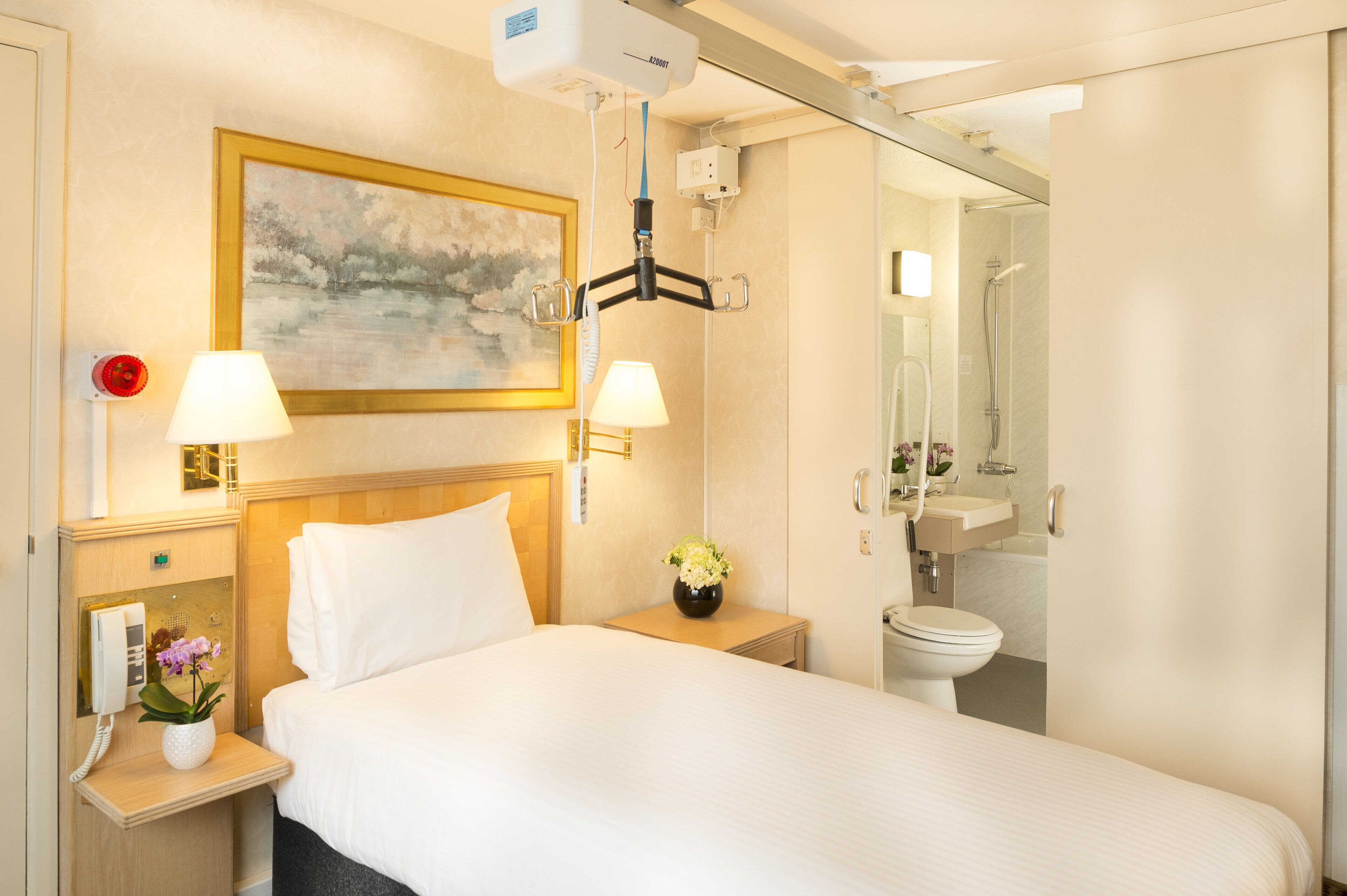 A hotel room with a bed, two bedside lamps, a painting, a vase with flowers, a telephone, and a bathroom with a shower and sink.