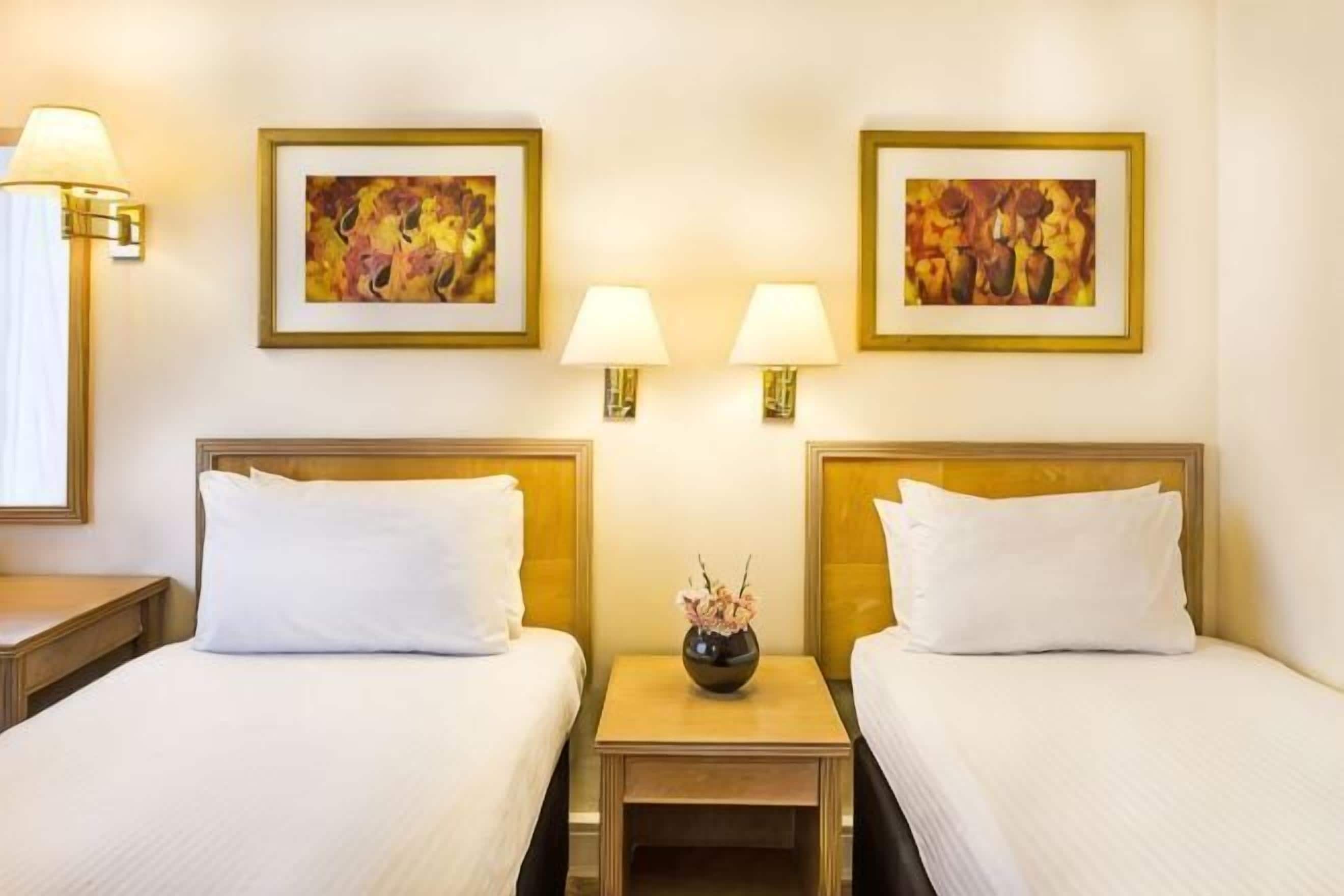 A hotel room with two beds, bedside lamps, and two framed artworks on the wall.