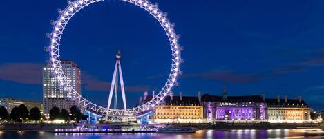 Une grande roue avec une tour centrale, entourée de bâtiments illuminés et bordée par une étendue d’eau.