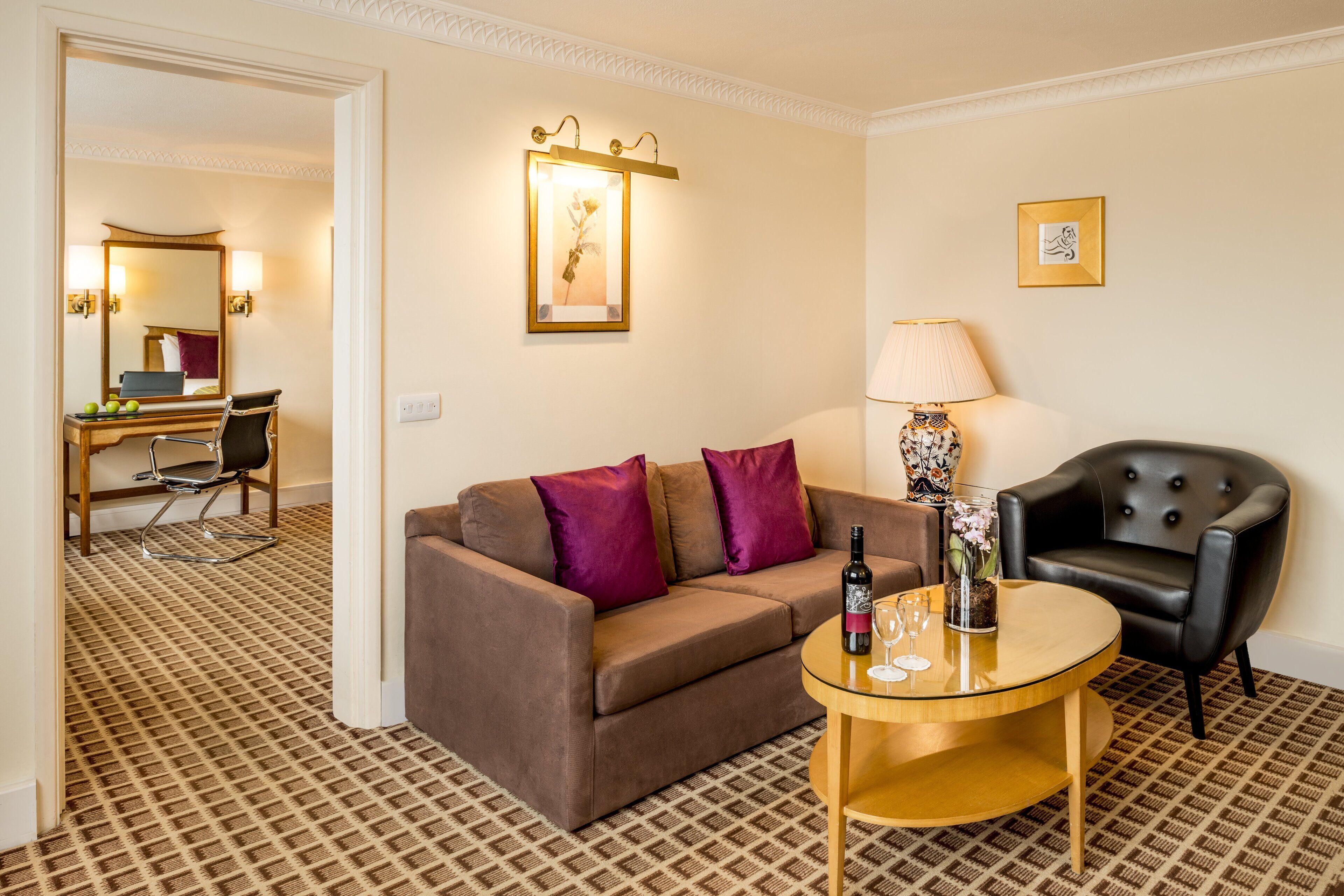 A hotel room with a brown sofa, a round coffee table with a bottle and glasses, a black armchair, a lamp, and a framed picture on the wall.
