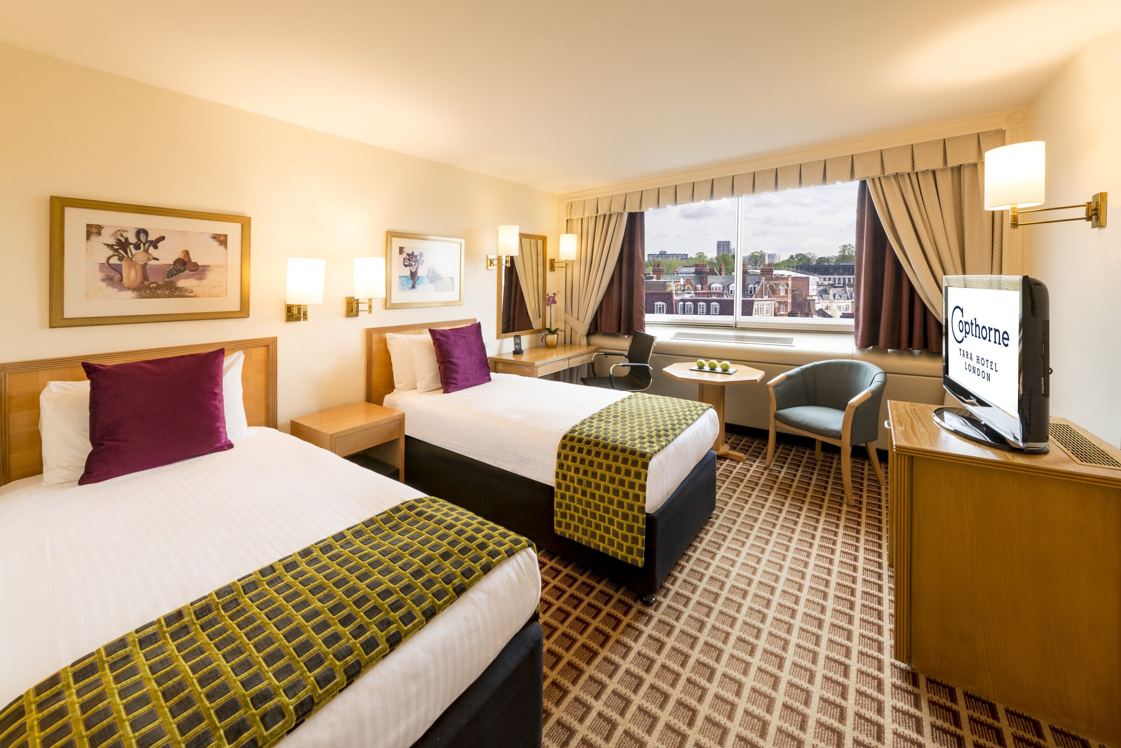 Ein Hotelzimmer mit zwei Betten, einem Schreibtisch, einem Sessel, einem Fernseher und einem Fenster mit Blick auf die Stadt.