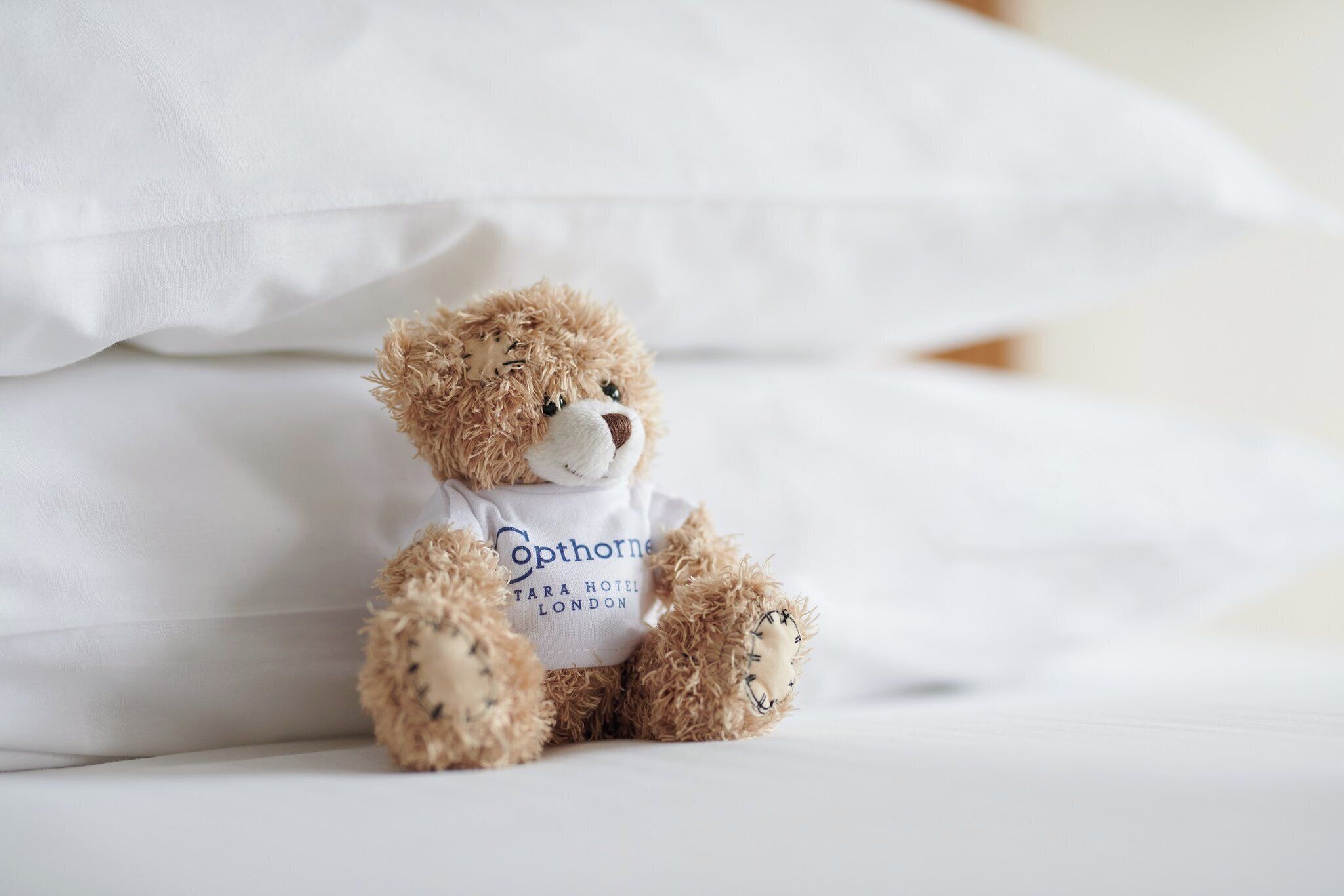 Un ours en peluche portant une chemise blanche avec l’inscription « Cophorne Hotel London », assis sur un lit recouvert de draps blancs.