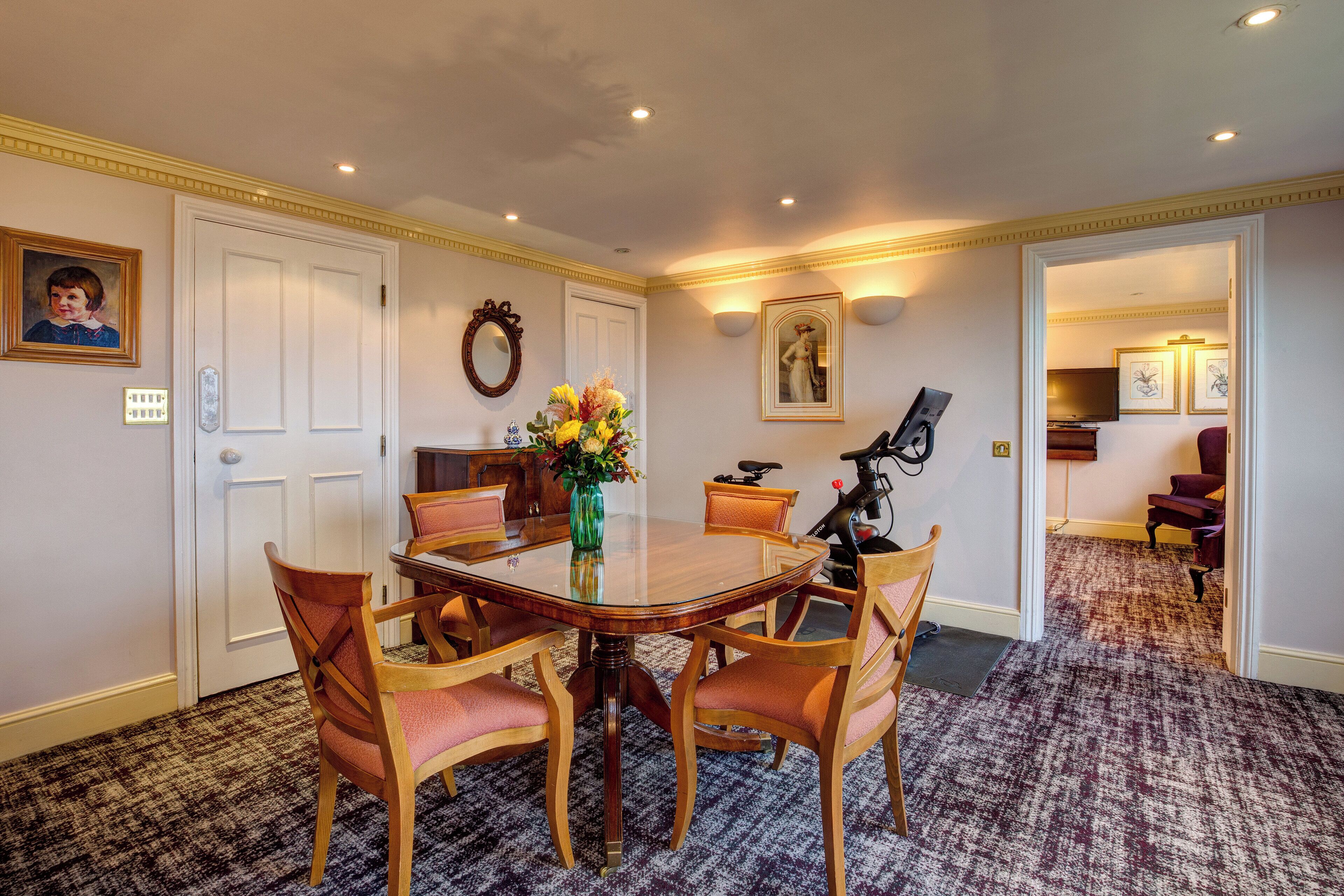 A dining room with a glass table surrounded by wooden chairs, a vase with flowers, and a painting on the wall.