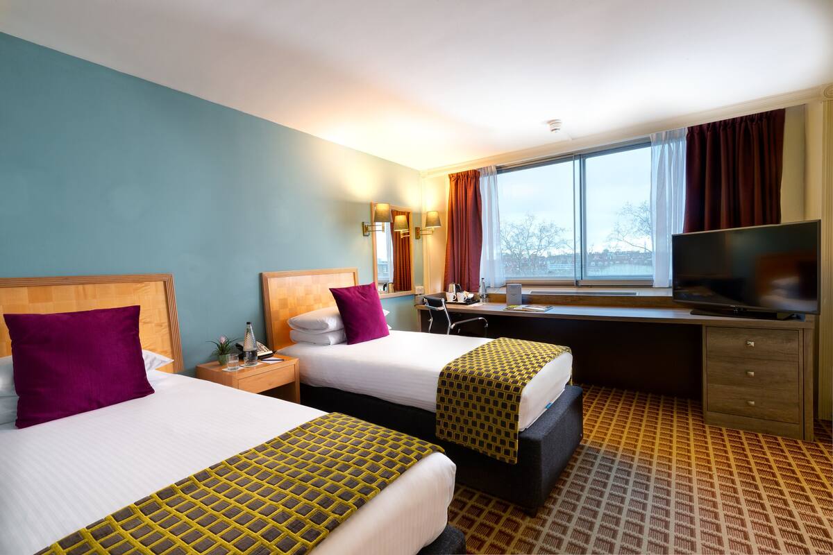 Ein Hotelzimmer mit zwei Betten, einem Schreibtisch, einem Fernseher und einem Fenster mit Vorhängen.