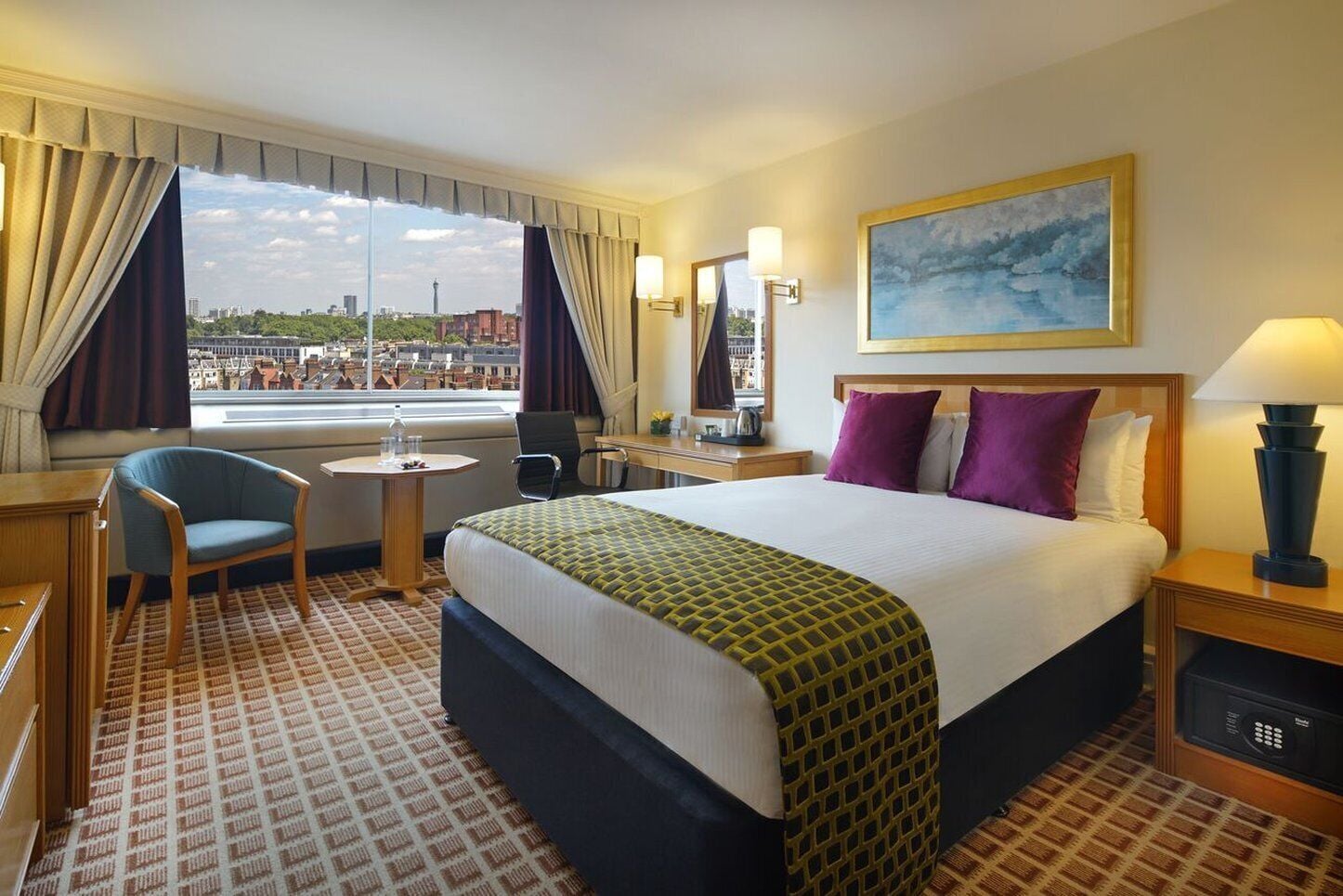 A hotel room with a large bed, a desk, a chair, a window with curtains, a painting on the wall, and a view of the cityscape.