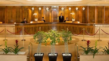 A hotel reception area with a wooden reception desk, receptionists, and a clock.