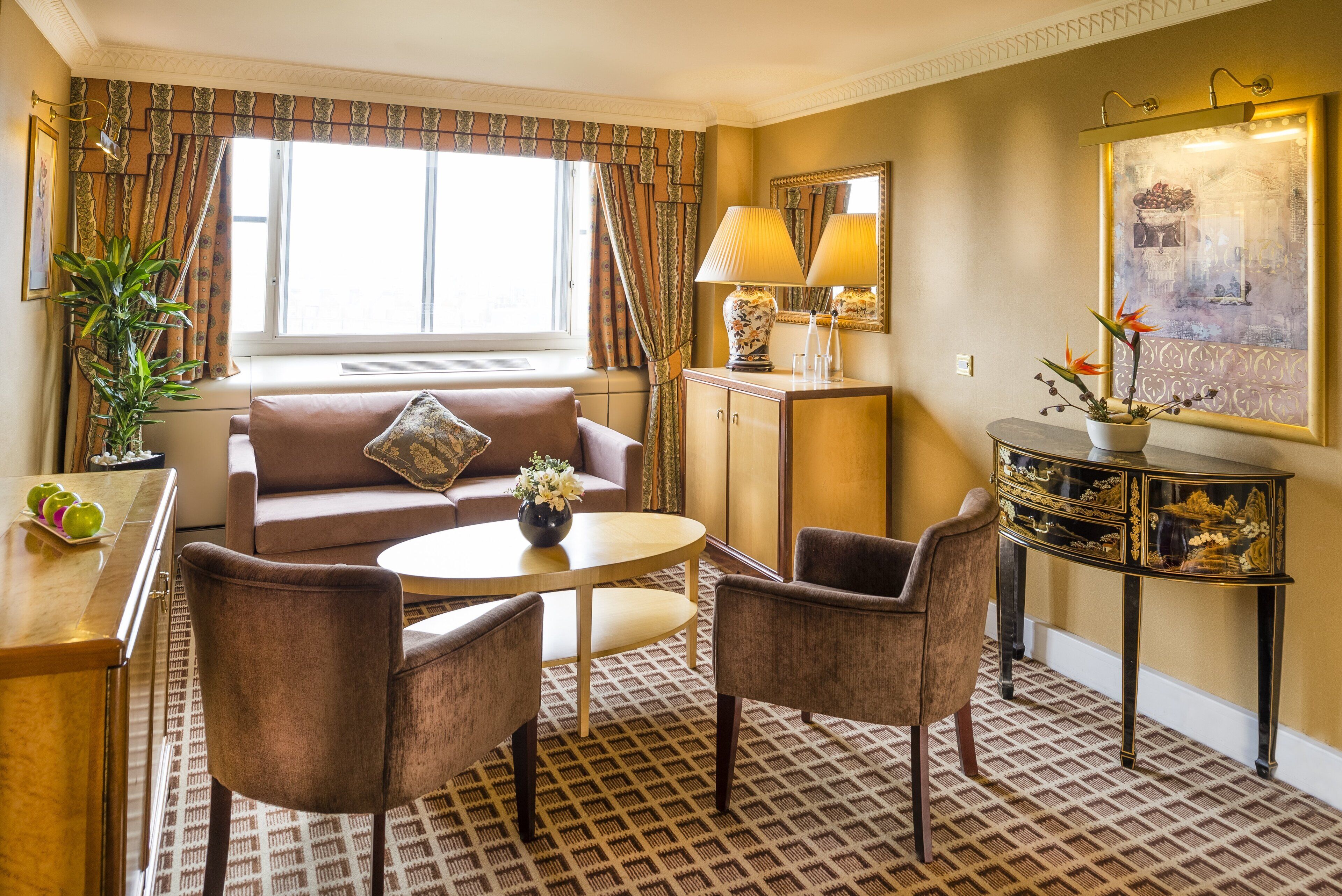 A hotel room with a sofa, two armchairs, a small round table, a cabinet, a potted plant, and a large window with curtains.