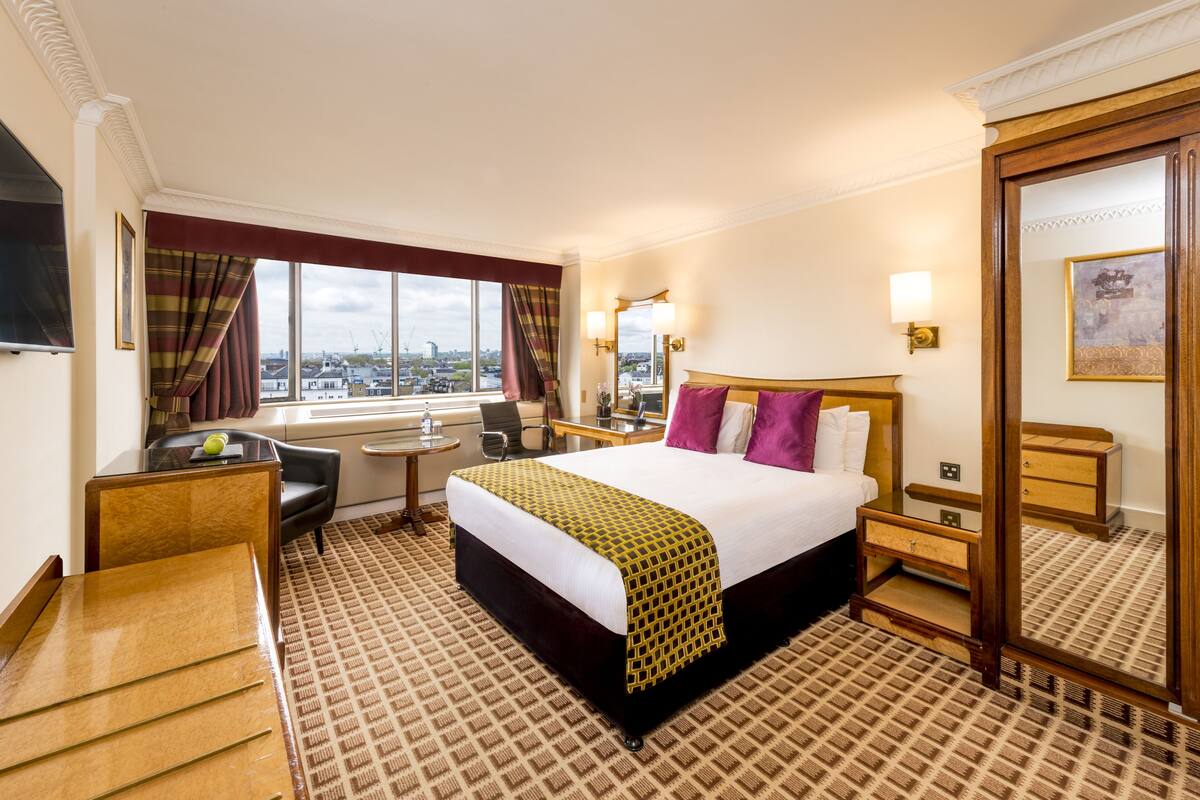 Ein Hotelzimmer mit einem großen Bett, einem Schreibtisch, einem Sessel, einem Fernseher und einem Fenster mit Vorhängen.