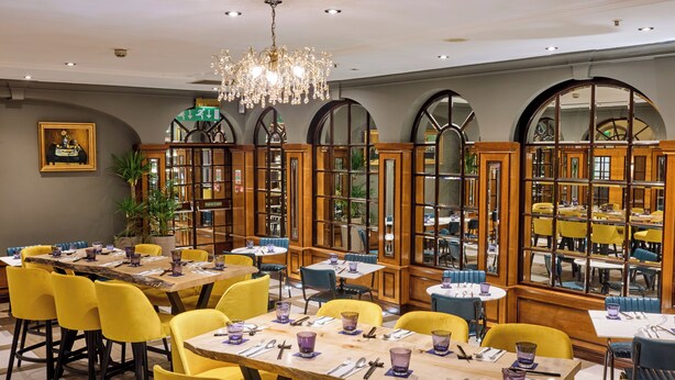 A restaurant with wooden tables and yellow chairs, a chandelier, and large windows with arched tops. - Copthorne Tara Hotel London Kensington (London)