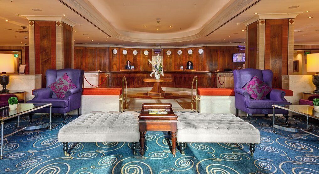 A lobby with a reception desk, two purple armchairs, a tufted ottoman, and a patterned carpet.