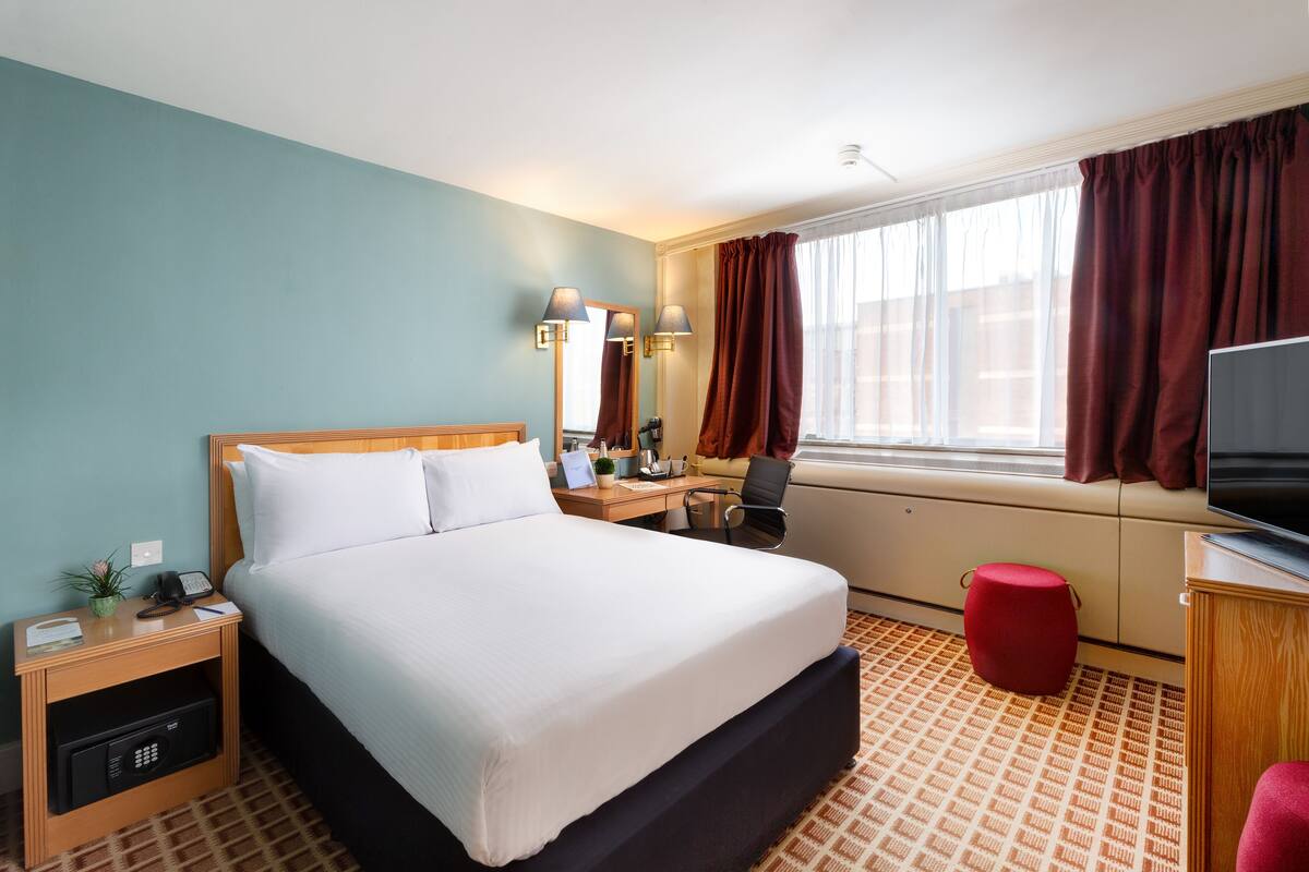Ein Hotelzimmer mit einem Bett, einem Schreibtisch mit Stuhl, einem Fernseher, einem Fenster mit Vorhängen und einem roten Hocker.