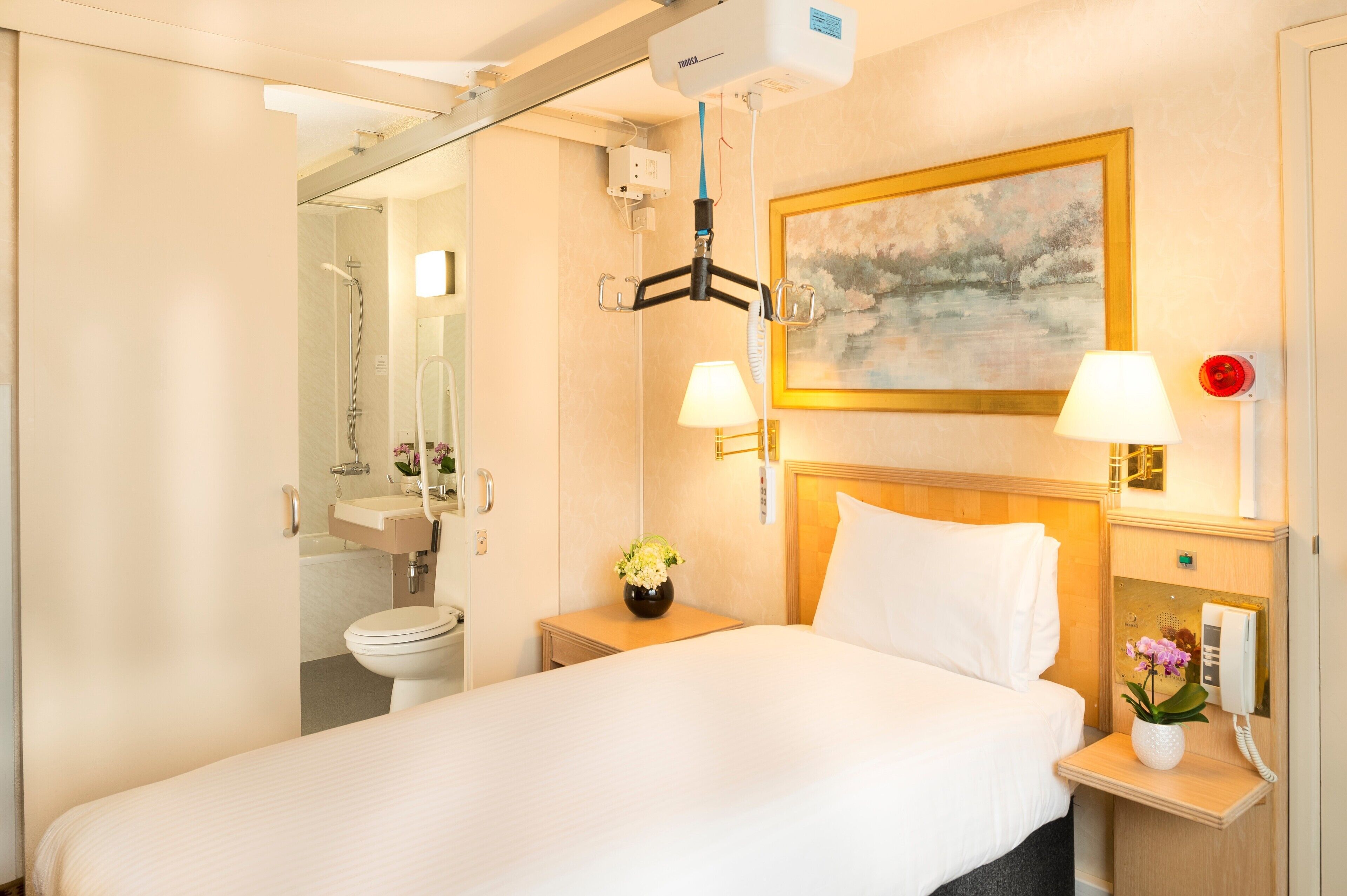 A hotel room with a bed, bedside lamps, a painting on the wall, a telephone, and a bathroom visible through an open door.