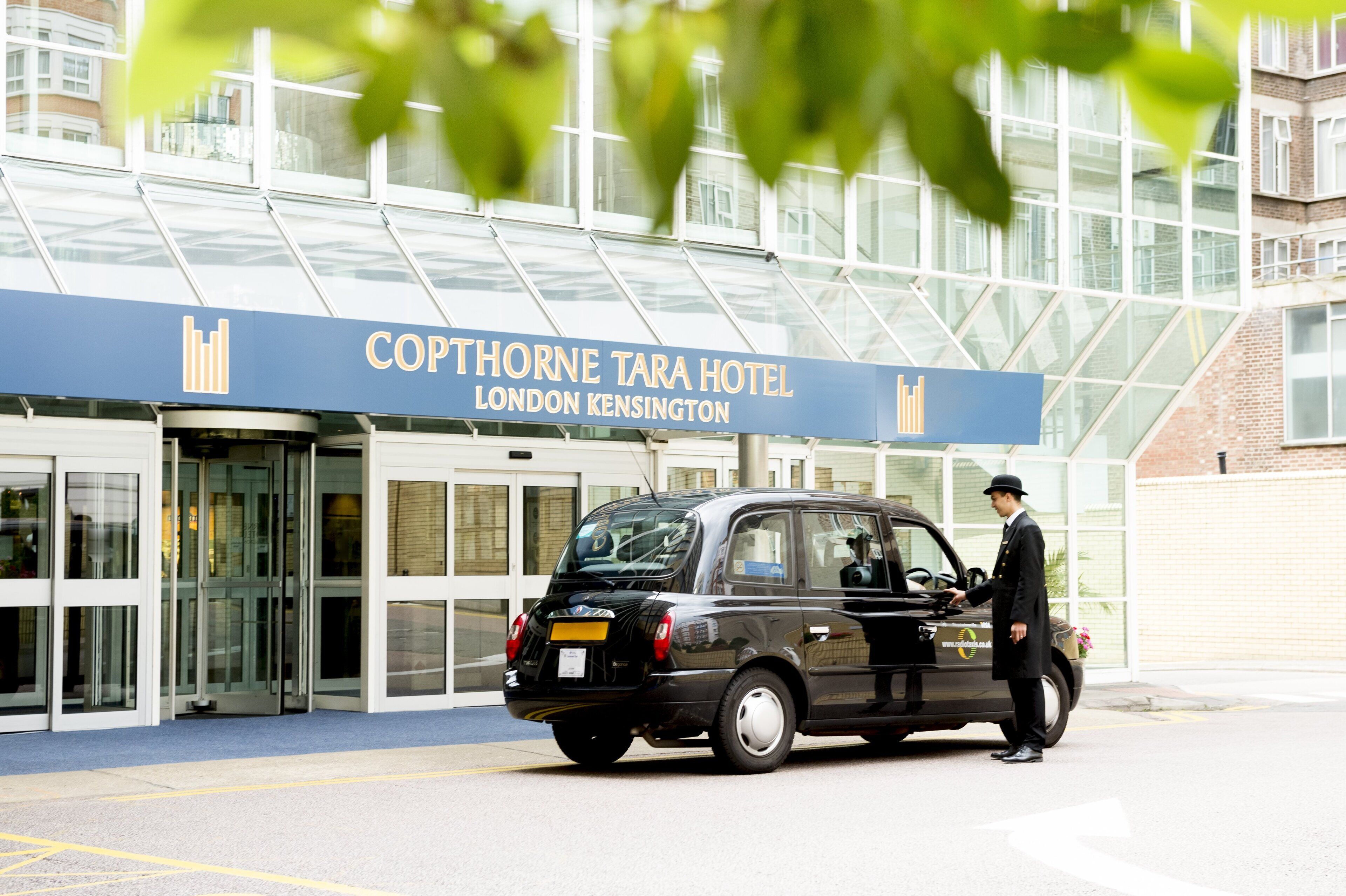 A black car parked in front of the Copthorne Tara Hotel, London Kensington, with a man in a suit standing beside it.