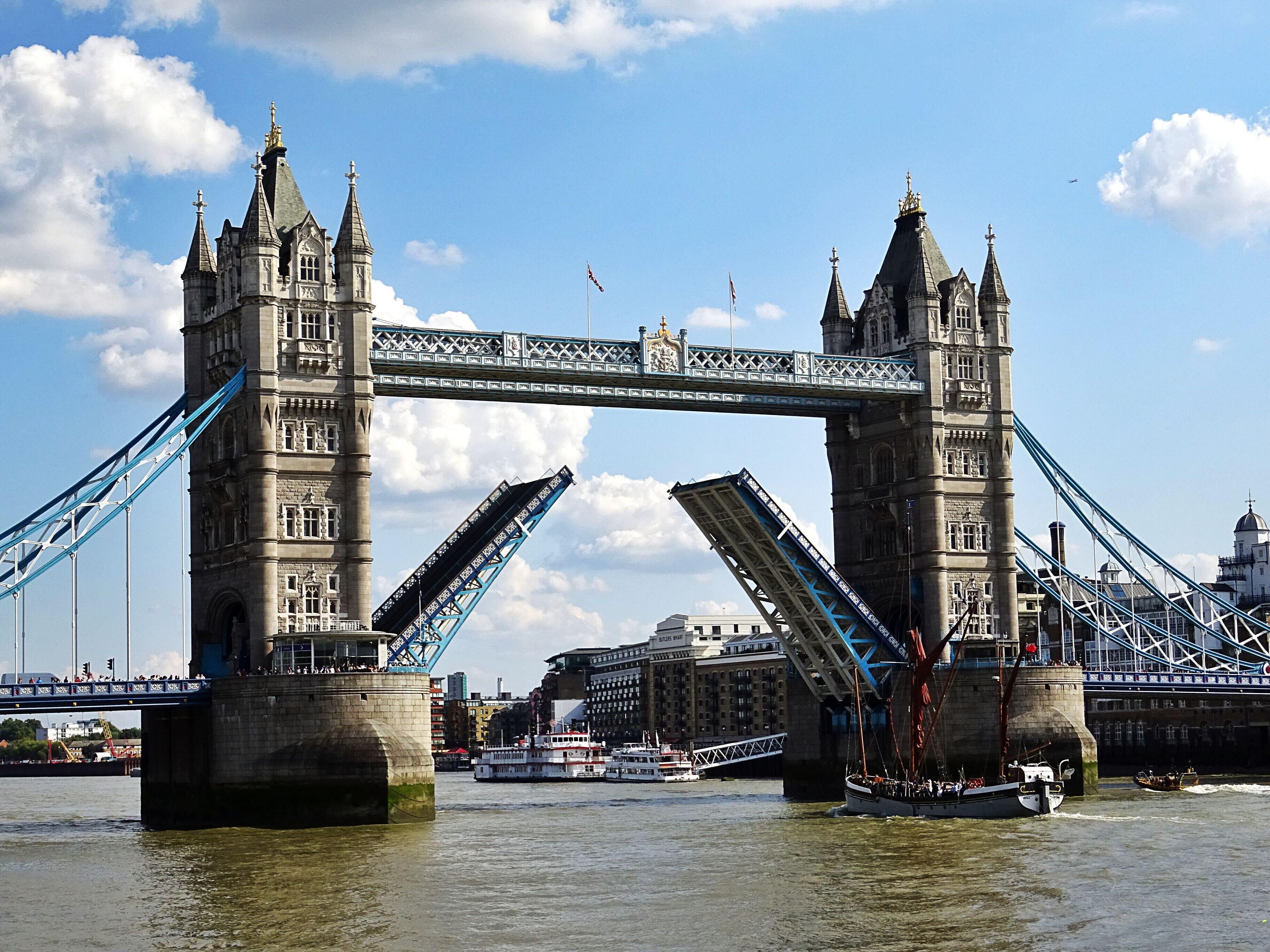 A historic bridge with two towers and a blue suspension section, spanning a river with boats and buildings in the background.