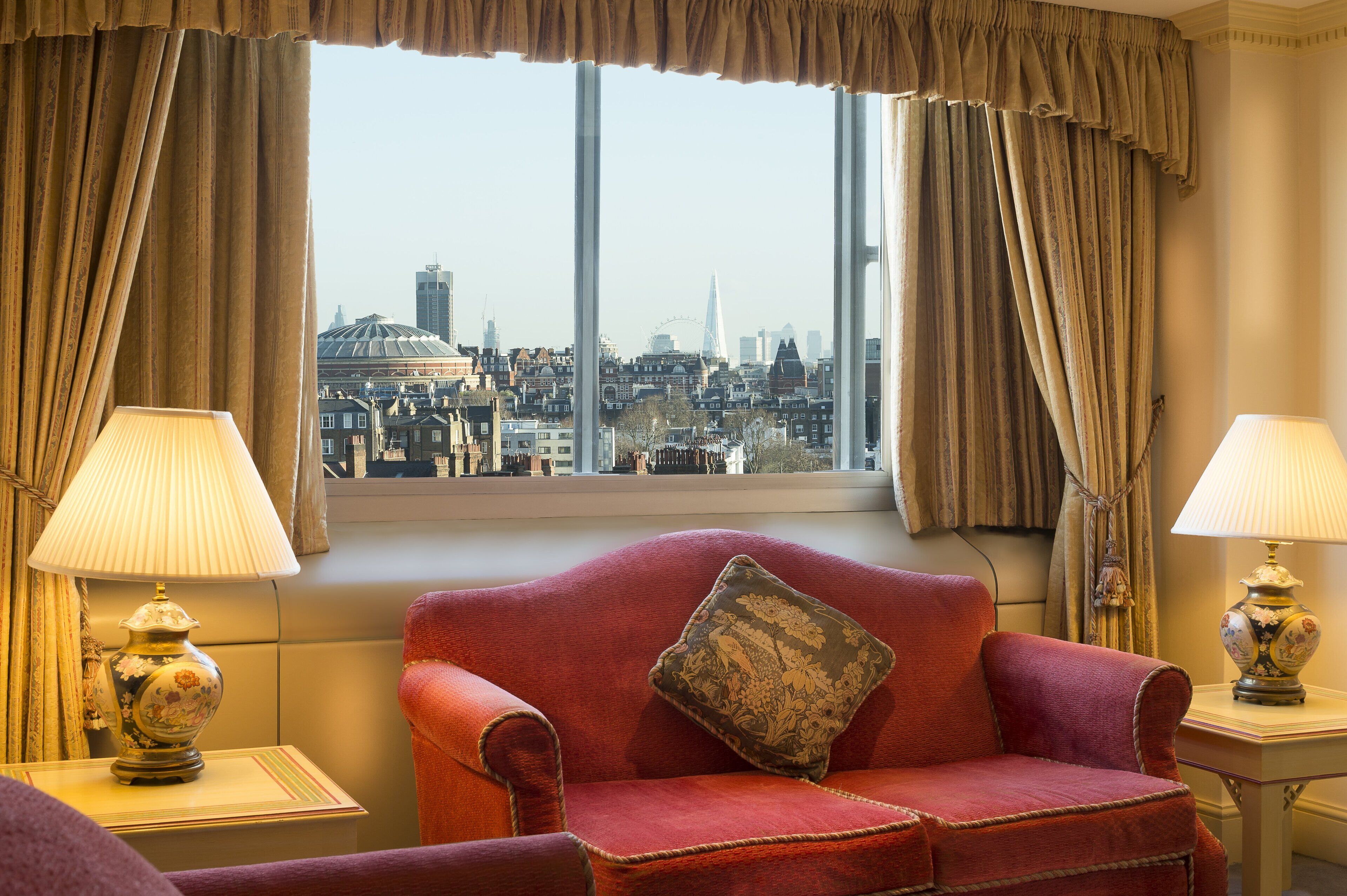 Una habitación con un sofá rojo, dos lámparas y vistas a un paisaje urbano a través de ventanas amplias.
