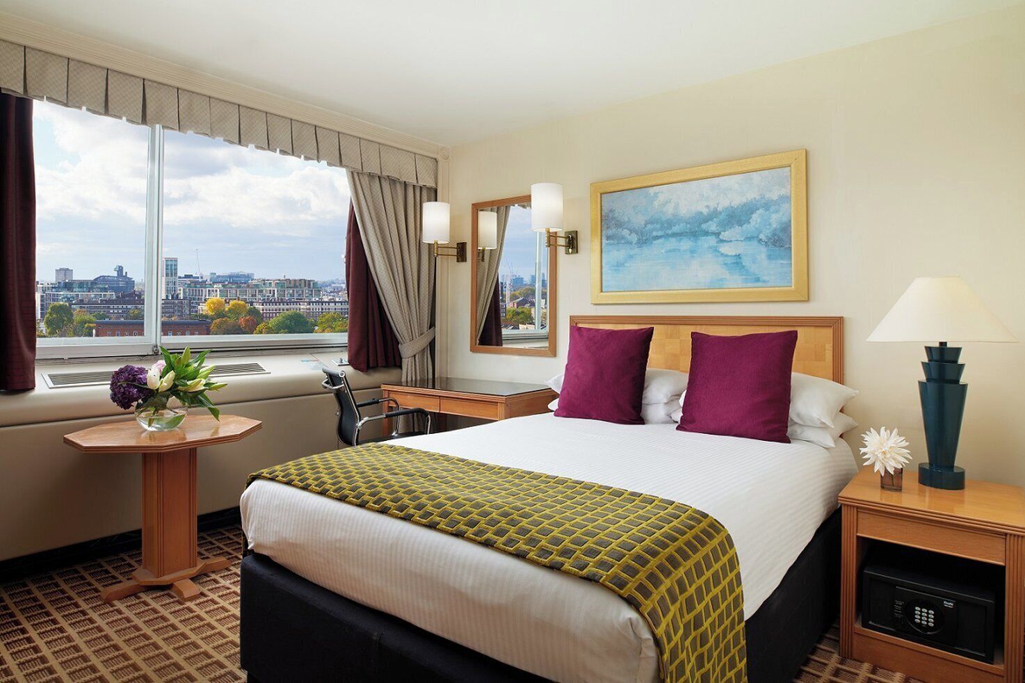 Een hotelkamer met een groot bed, een bureau, een stoel, een raam met uitzicht op de stad, een schilderij aan de muur, en een nachtkastje met een lamp en een bloemenvaas.
