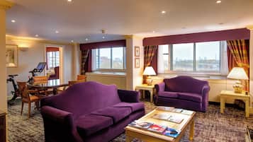 Une chambre d’hôtel avec un canapé violet, une table basse avec des magazines, un coin repas avec une table et des chaises, et de grandes fenêtres avec des rideaux.