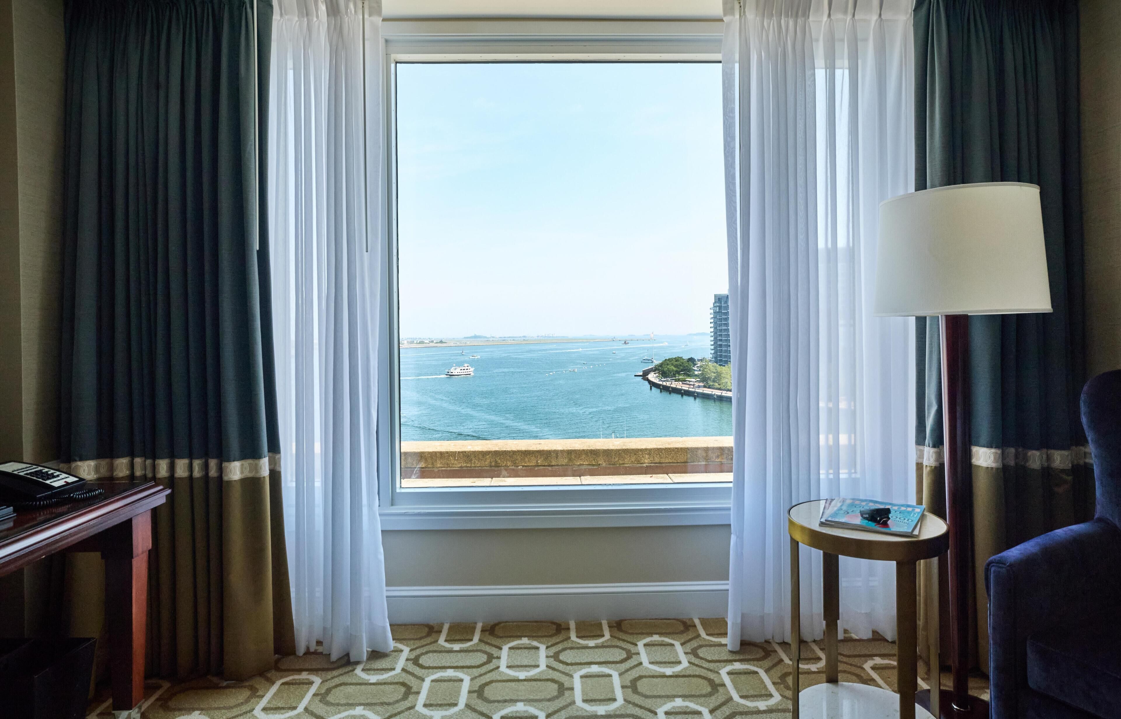 A hotel room with a large window, a desk, a chair, and a lamp.