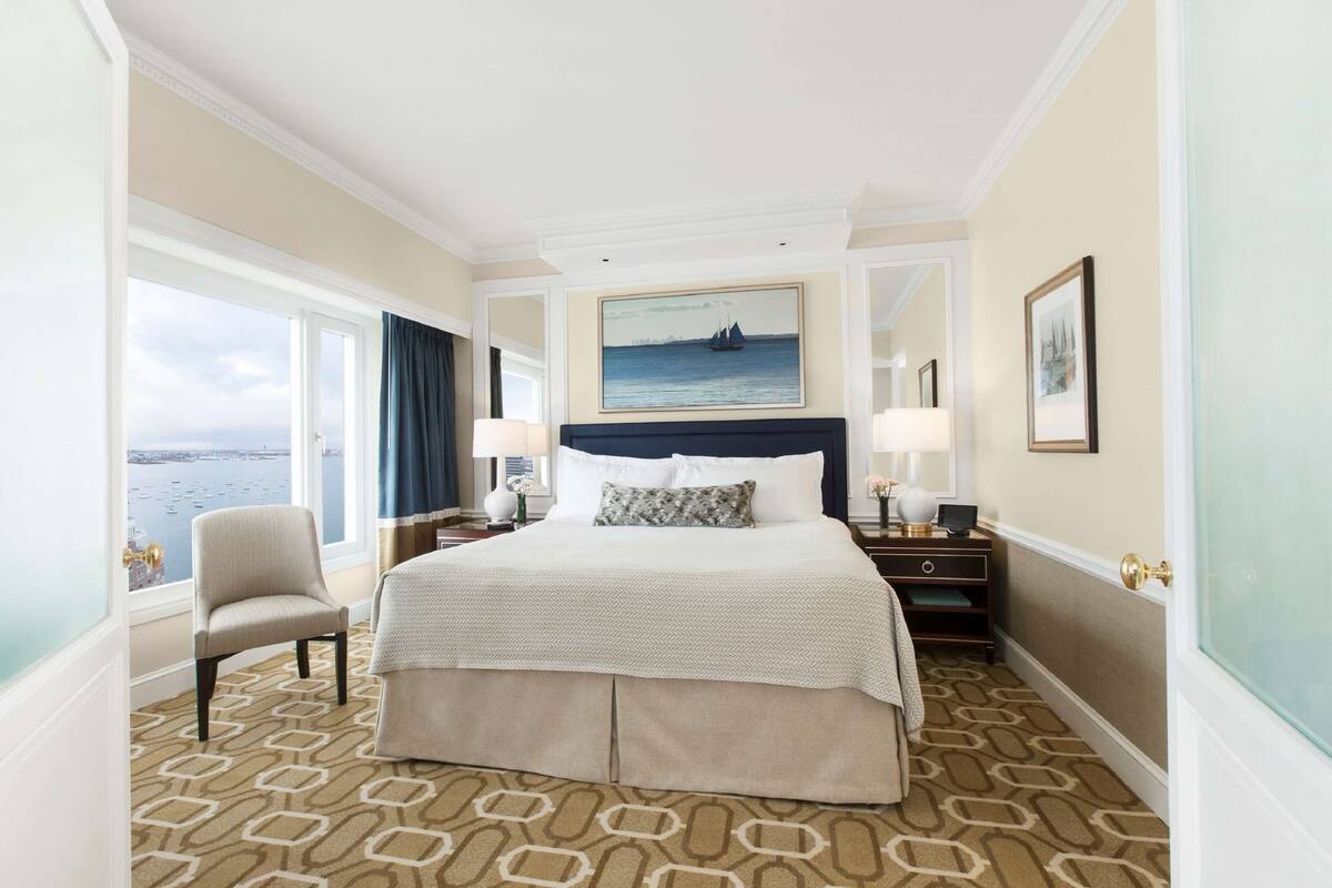 A hotel room with a large bed, a chair, a nightstand, a lamp, a painting, and a view of the sea.