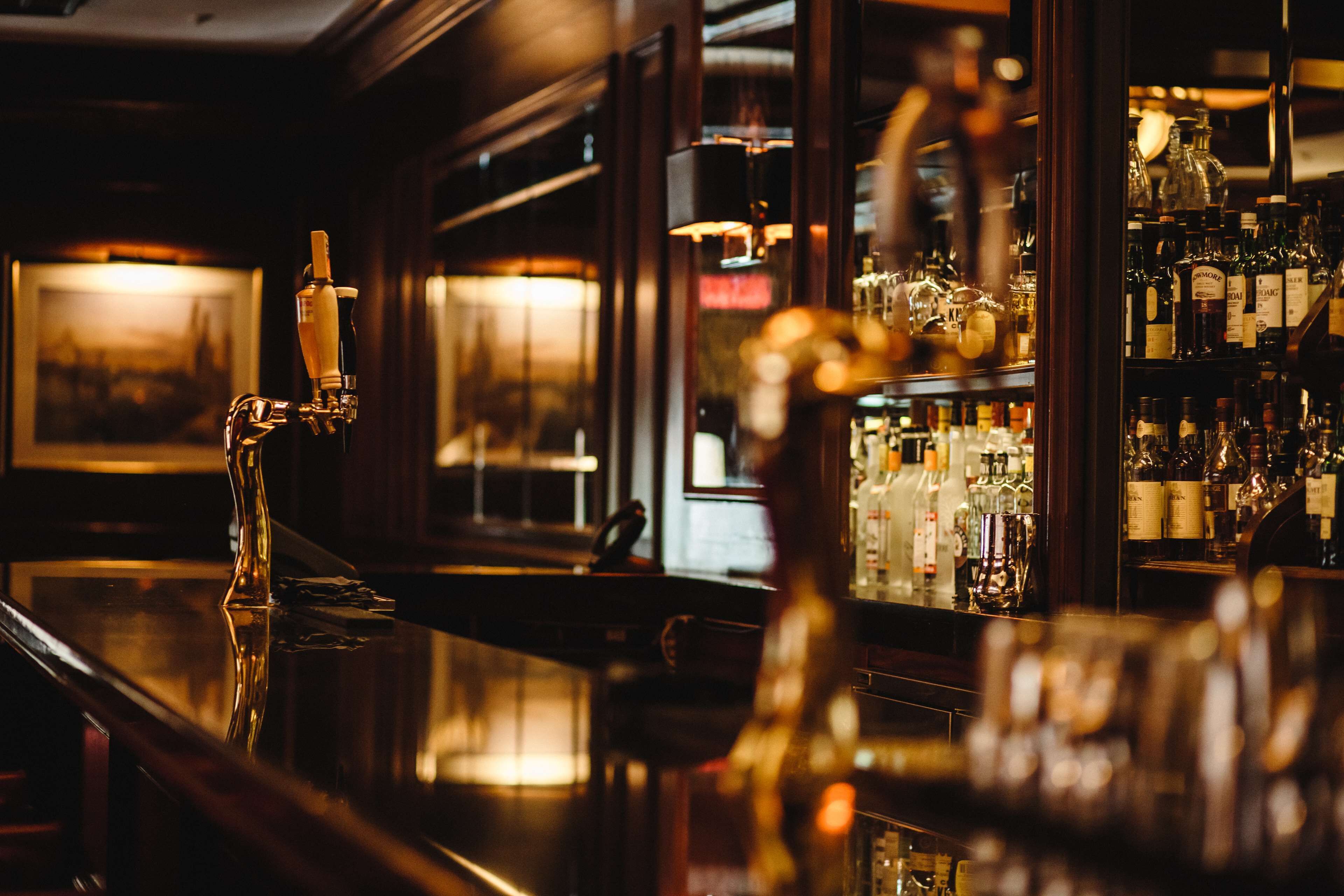 A bar with a polished wooden counter, a tap, and shelves stocked with bottles.